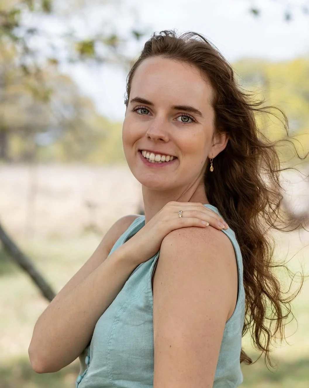 A woman with long, wavy brown hair smiling outdoors, wearing a sleeveless light blue top and jewelry, with a blurred background of trees and sky.