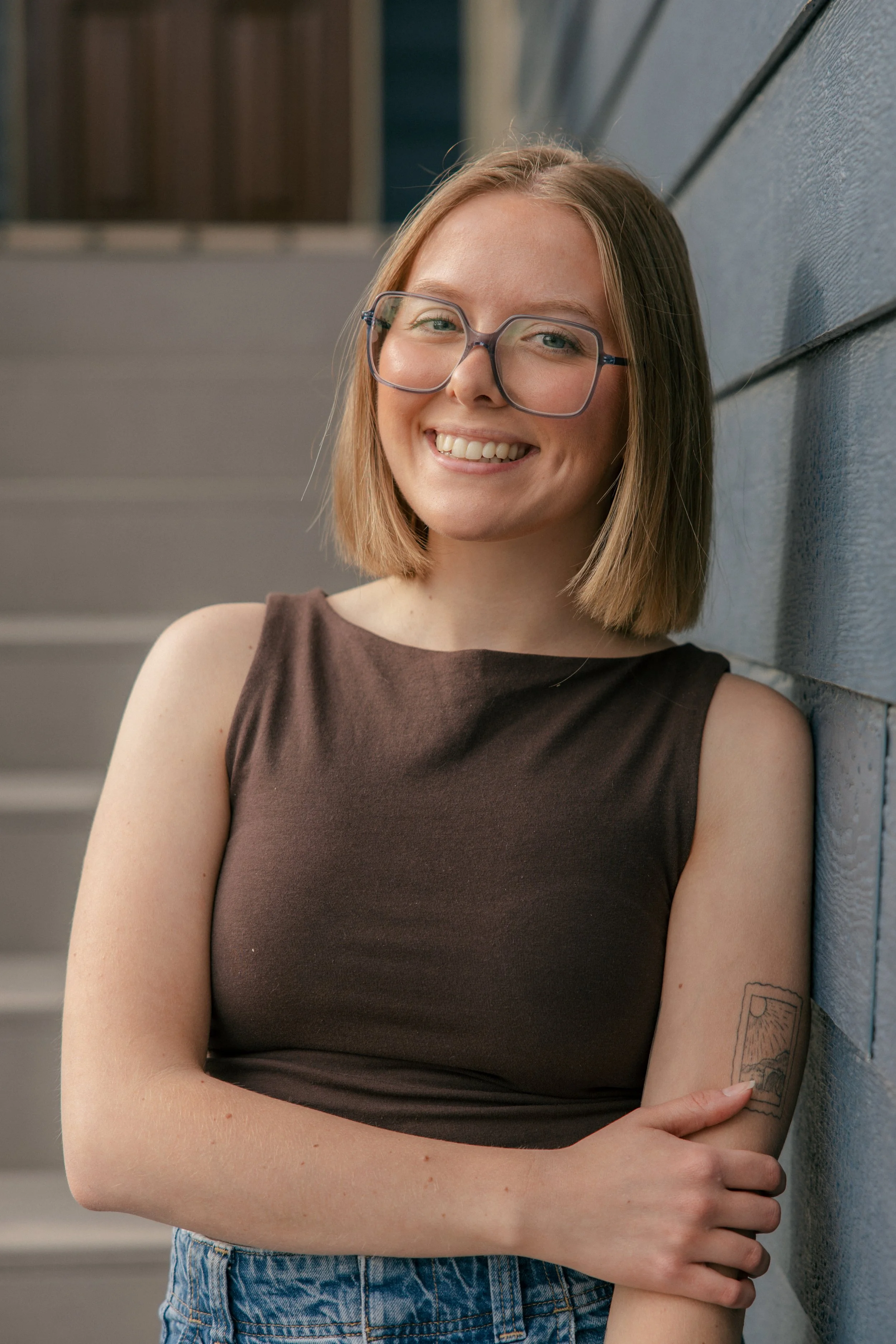 A young woman with shoulder-length brown hair, wearing glasses and a sleeveless brown top, standing against a gray wooden wall, smiling and looking at the camera.