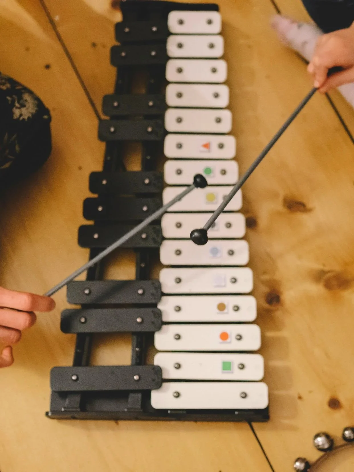A children's toy xylophone with white and black keys, some keys have colored dots or markings. Two hands are holding mallets, ready to play the instrument on a wooden table.