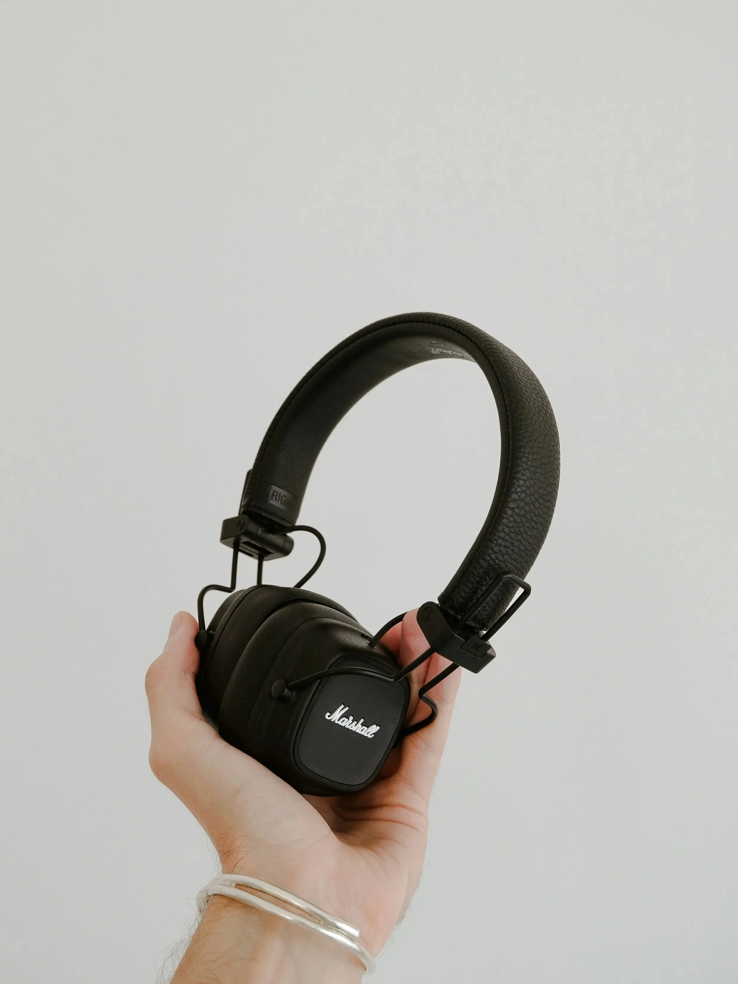 A person holding a black Marshall over-ear headphone with a white wall background.
