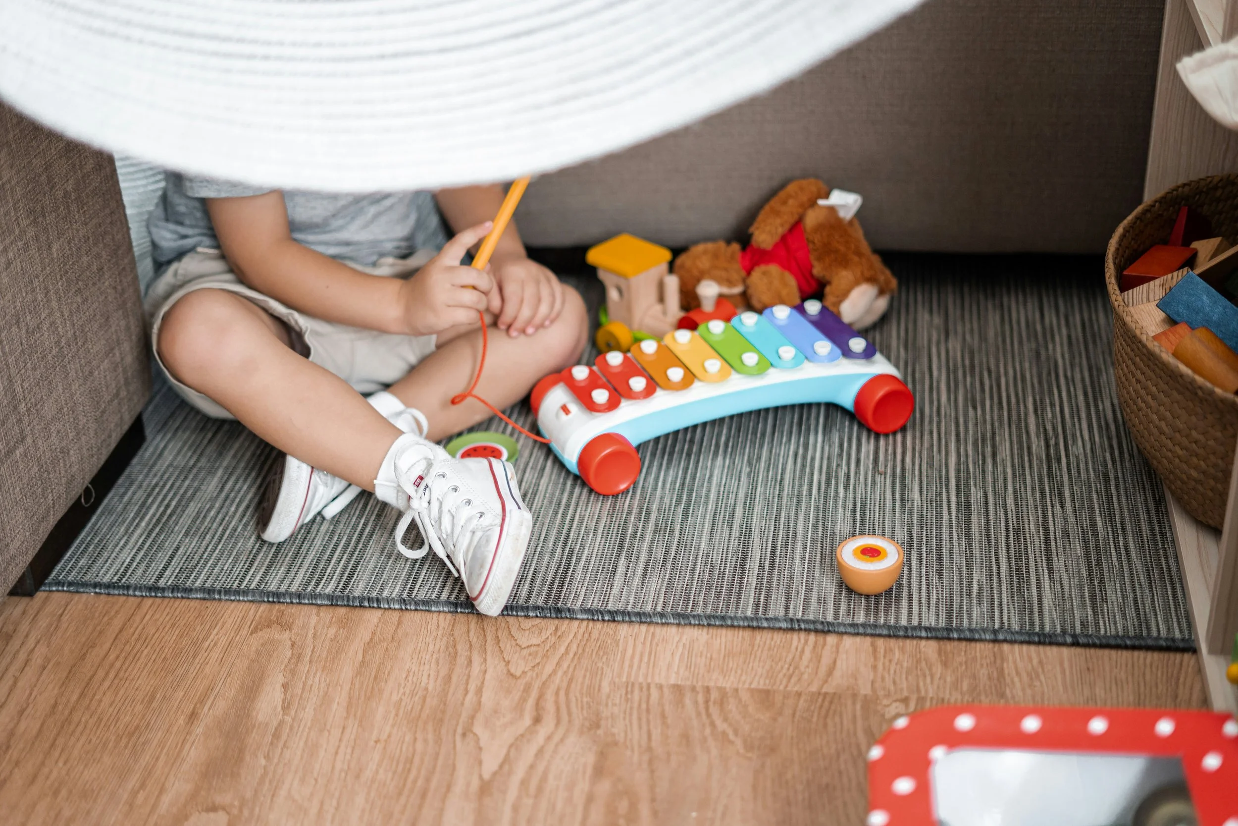 Child sitting on the floor playing with a colorful xylophone toy, with a teddy bear nearby. The photo is taken from above, partially obscured by a large white object.