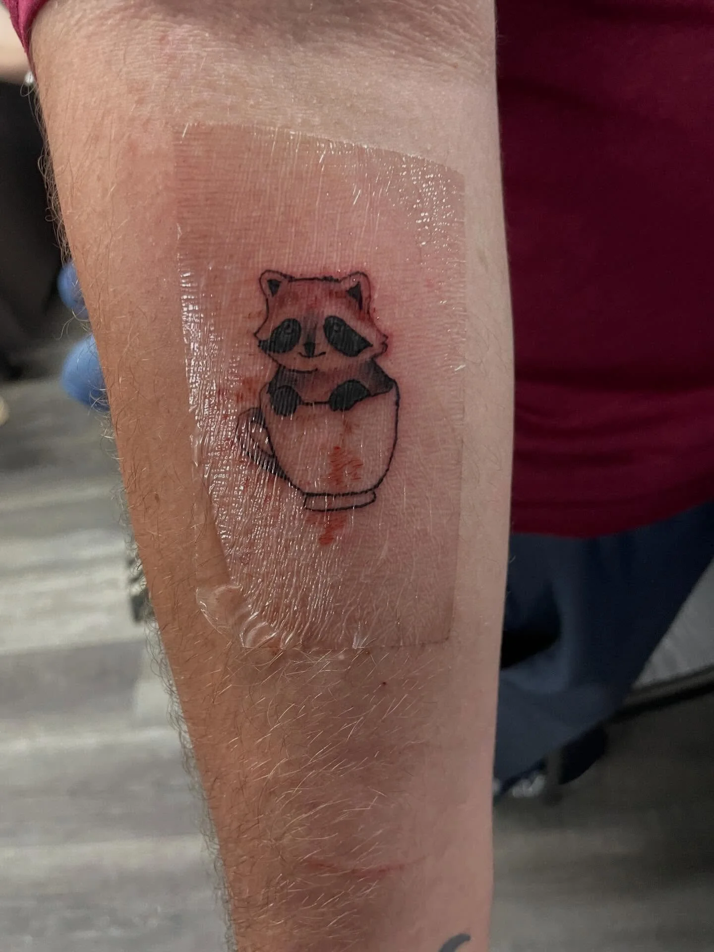 Temporary tattoo of a cute raccoon holding a jar on a person's forearm, covered with clear protective tape.