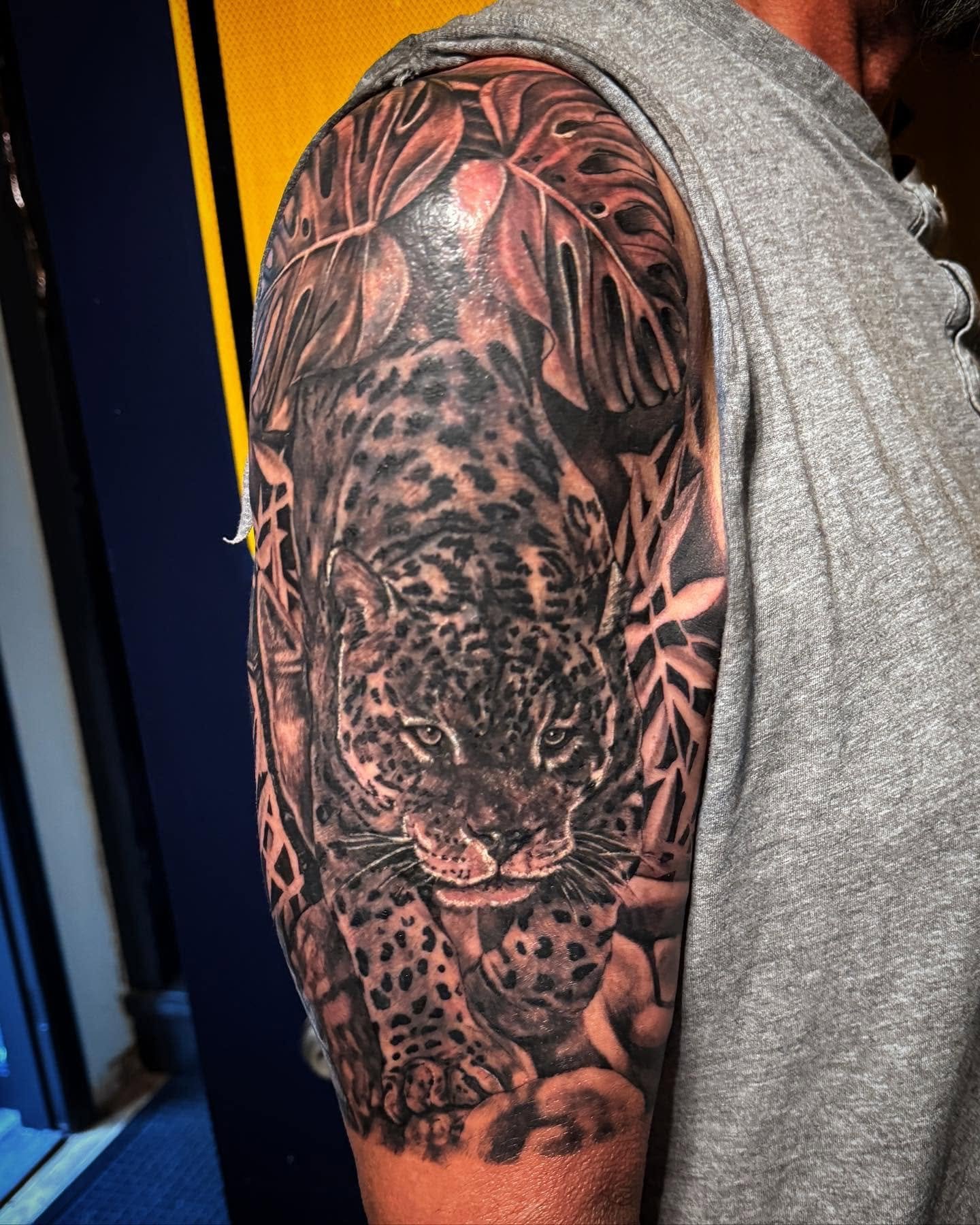 Tattoo of a realistic jaguar's face and body on a person's upper arm and shoulder, with leaves and tribal patterns in the tattoo design.