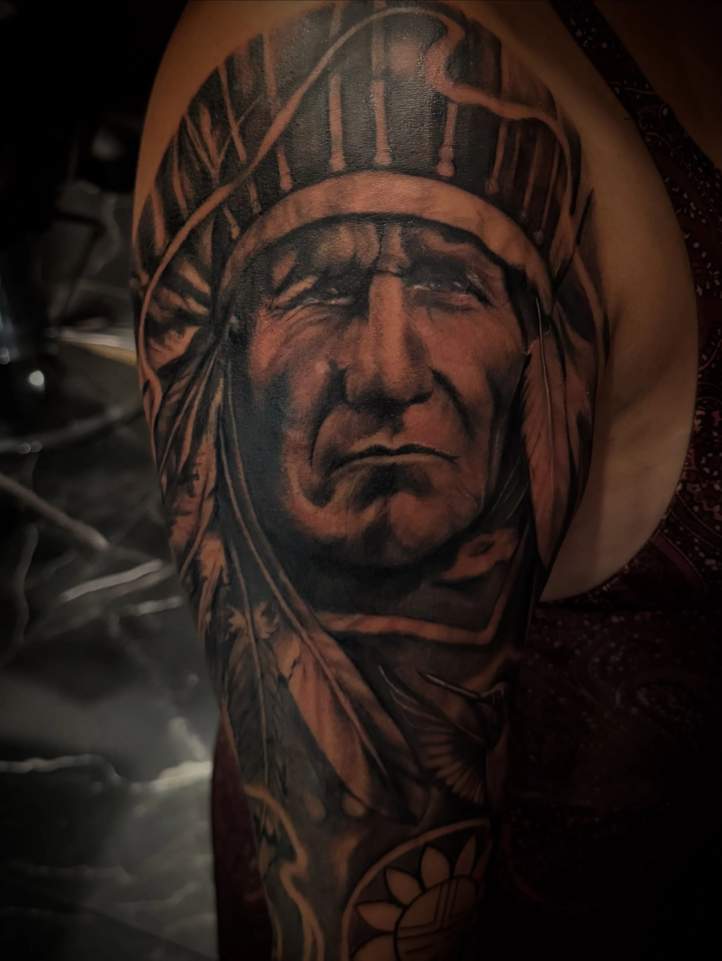 Close-up of a person's upper arm and shoulder tattoo featuring a Native American chief wearing a feathered headdress, with Roman numerals in the background.