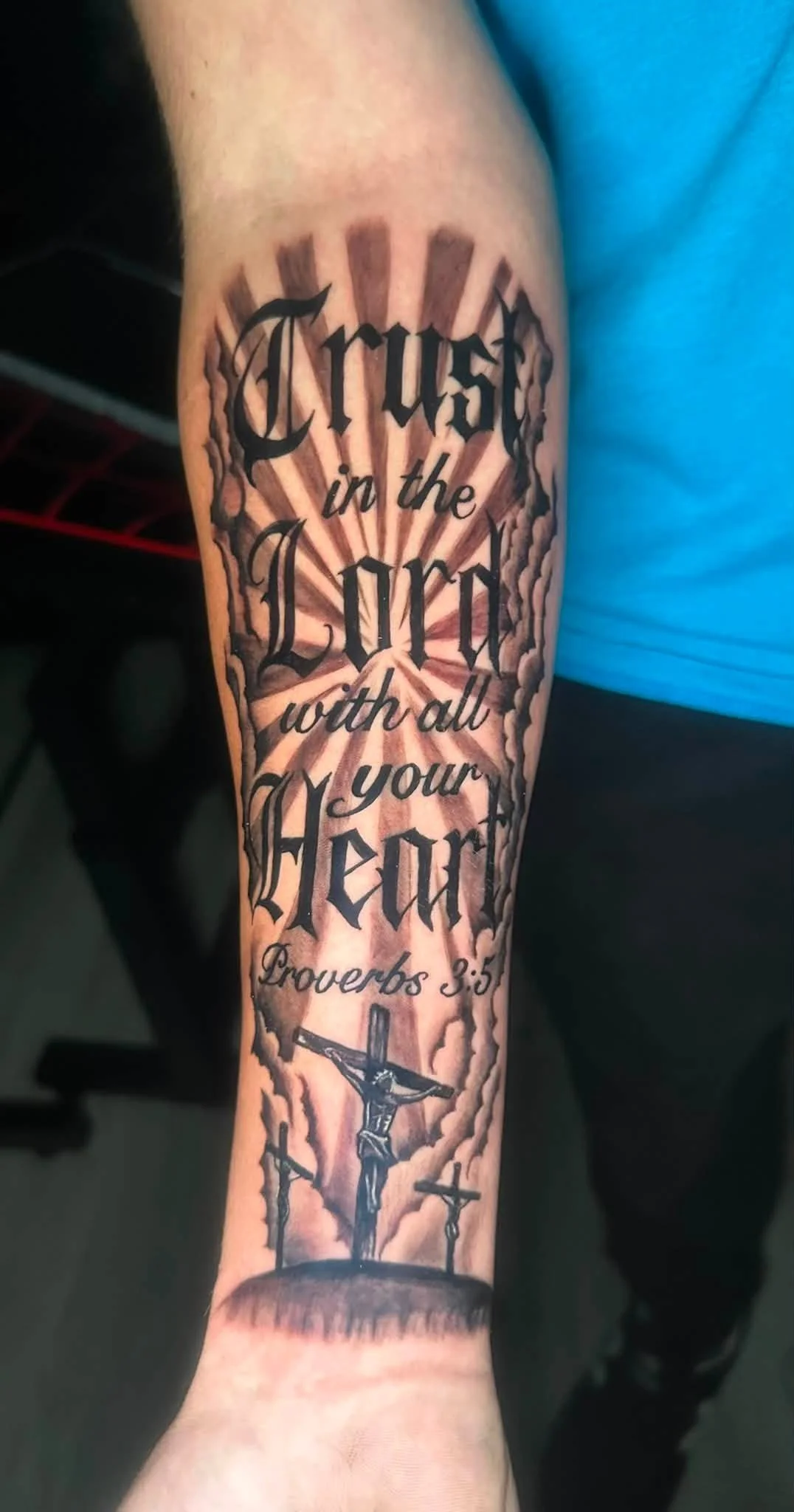 Tattoo on person's forearm featuring a Bible quote from Proverbs 3:5, with a depiction of Jesus crucifixion at the bottom.