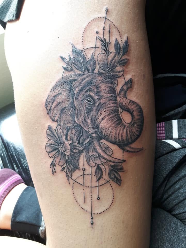 Tattoo of an elephant head surrounded by flowers and leaves, with geometric and dotted circle patterns above and below.