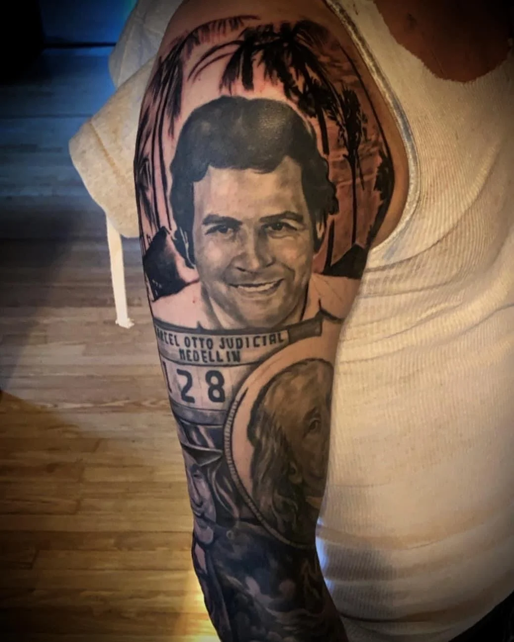 Tattoo of a smiling man's face on person's upper arm, with a background of a sunset and palm trees, and a tattoo of a woman’s face on the lower arm.