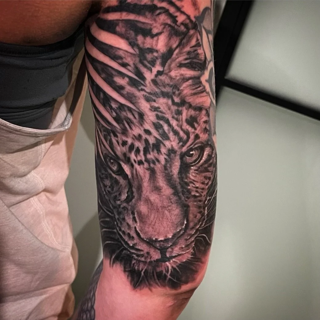 Close-up of a tattoo on a person's arm depicting a realistic black and gray lion's face with detailed shading and fur texture.