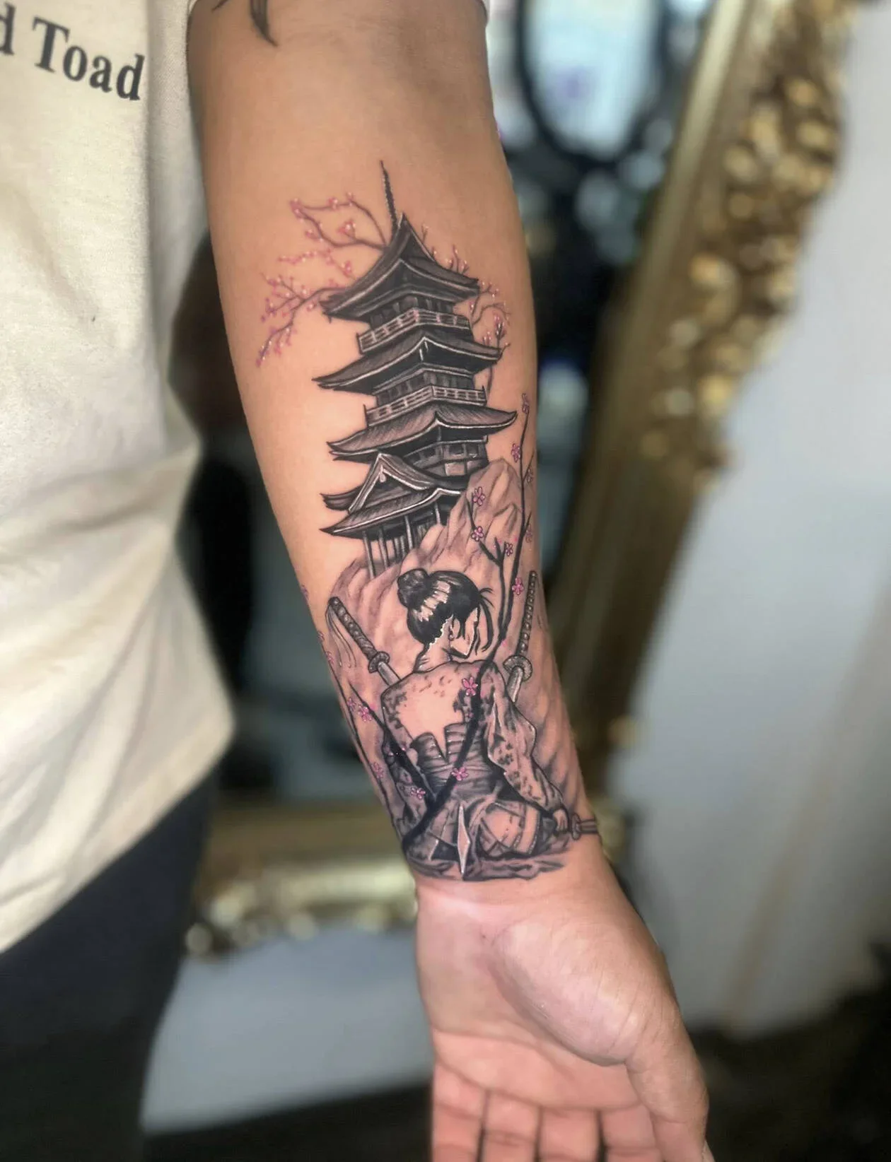 Tattoo of a traditional Japanese pagoda with cherry blossoms, depicting a woman in a kimono sitting with swords on her back.