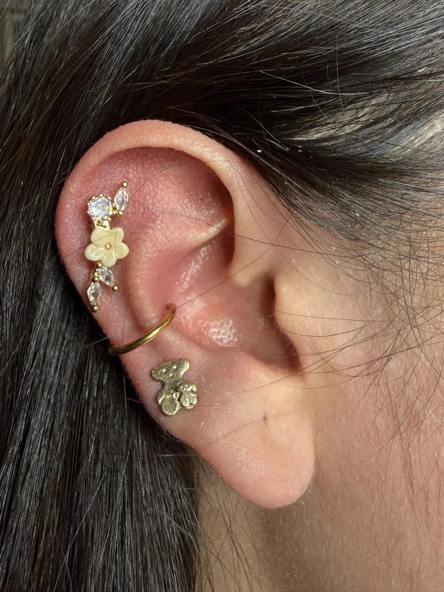 Close-up of a person's ear decorated with multiple gold and silver earrings, including a flower-shaped earring, a star-shaped earring, a round rhinestone, and small gold bars, with dark hair surrounding the ear.