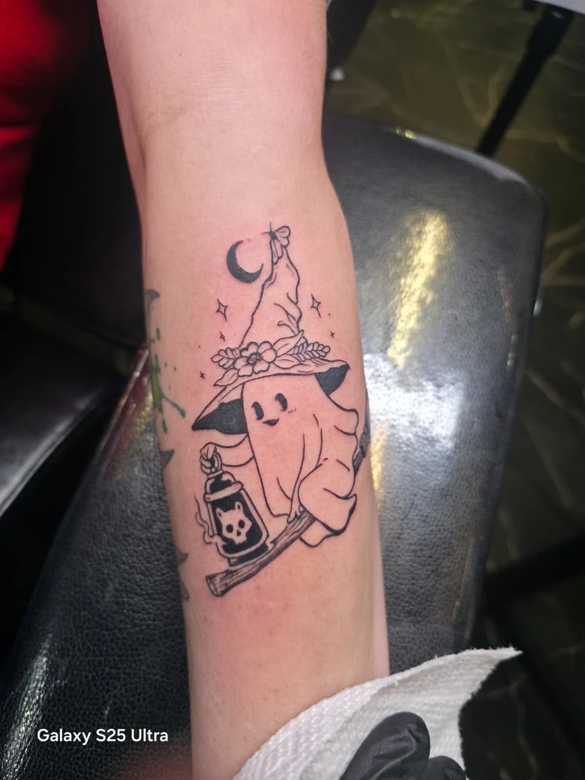 Black outline tattoo of a mushroom with a face, wearing a witch hat decorated with flowers, sitting on a branch holding a lantern with a skull design, against a crescent moon and starry sky background.