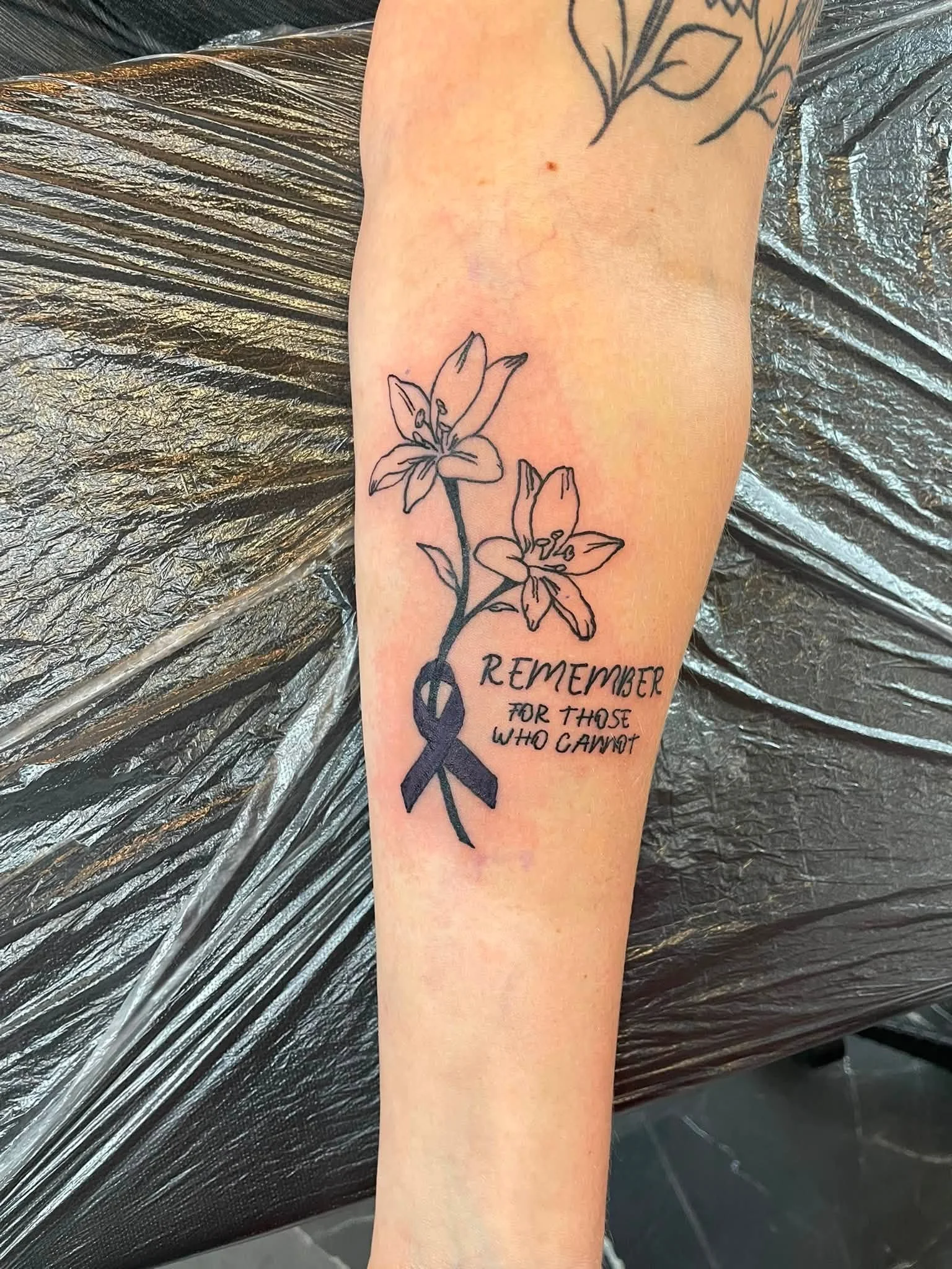 A tattoo of two lilies on an arm with a ribbon and the text 'REMEMBER FOR THOSE WHO CANNOT'.