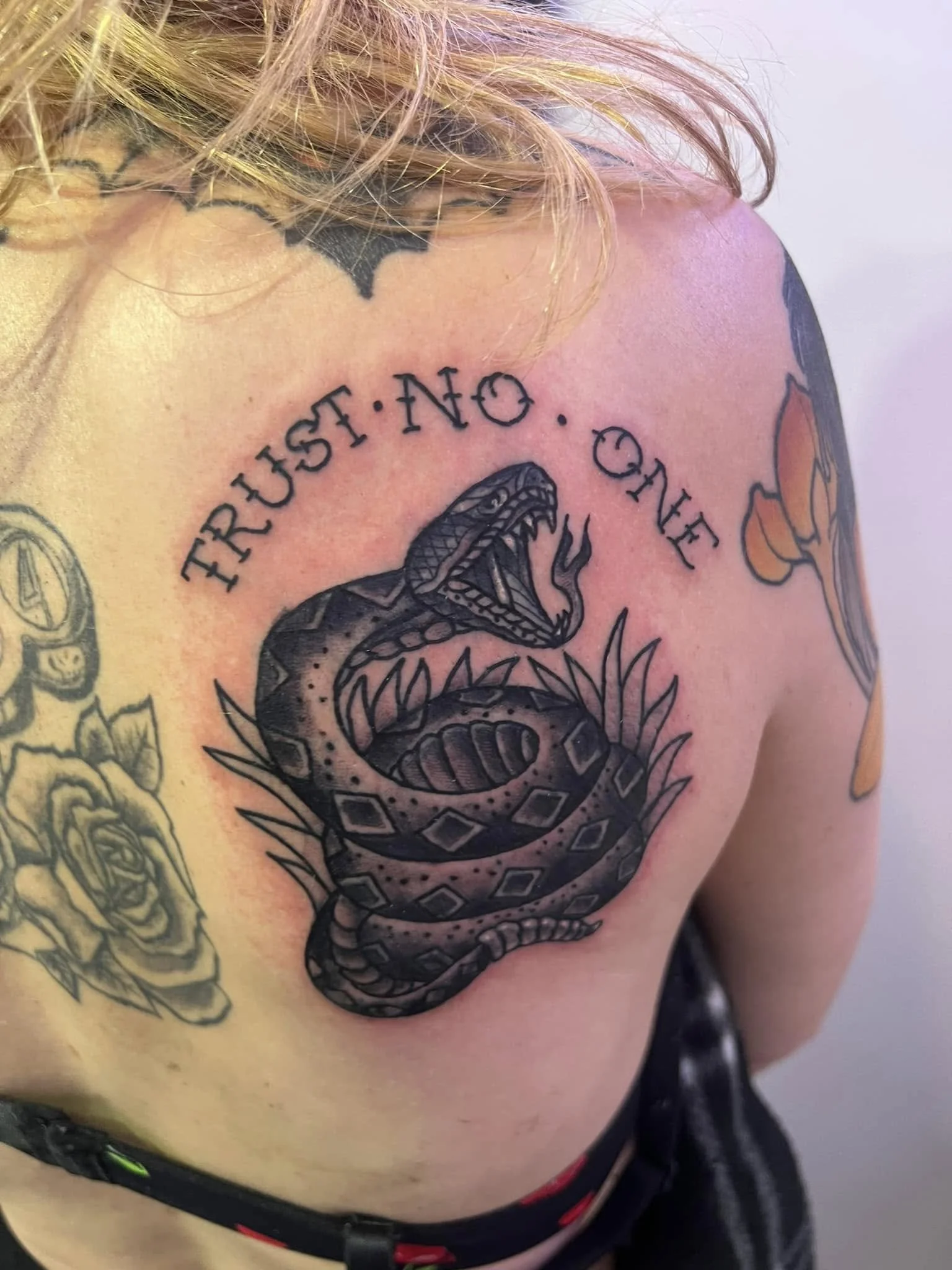 Close-up of a person's shoulder and upper chest displaying multiple tattoos, including a snake, a flower, and the phrase "Trust No One." The person has blonde hair.