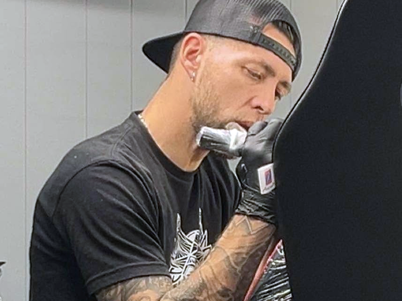 A man with tattoos, wearing a black baseball cap backwards and black gloves, is leaning against a black surface, looking down at a smartphone.