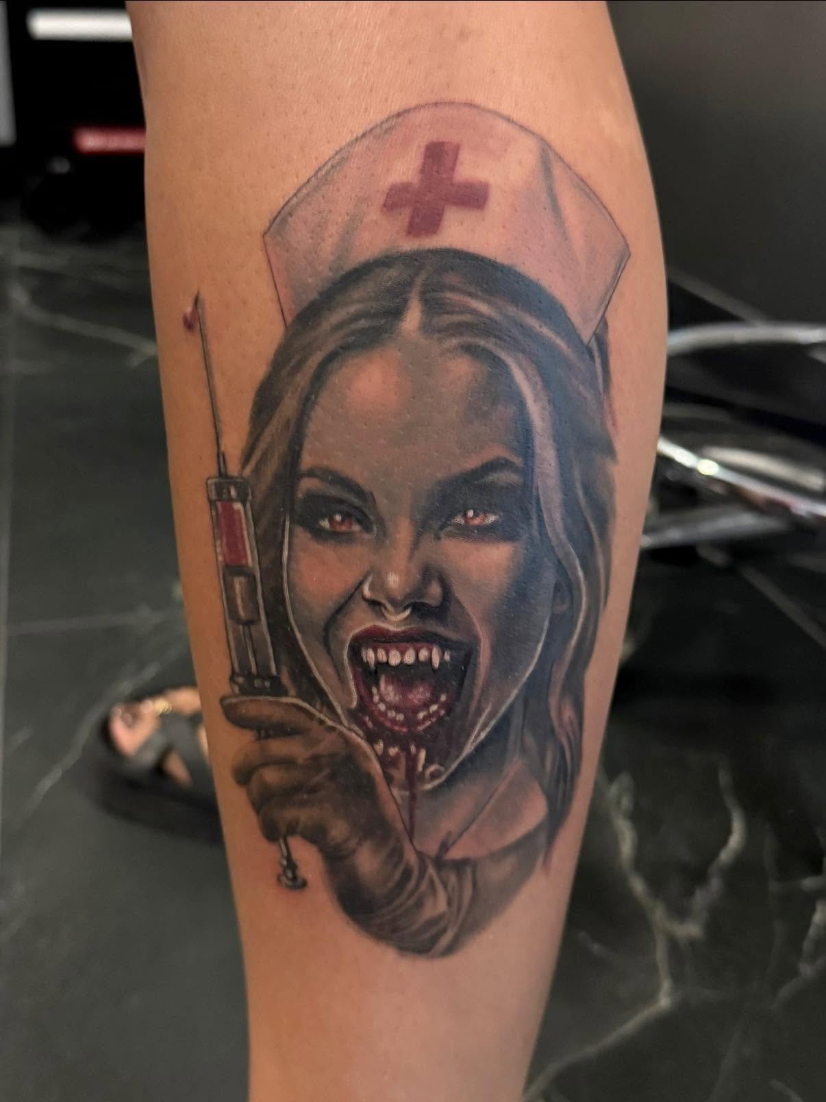 Tattoo of a woman with vampire-like teeth wearing a nurse's hat with a red cross, smiling with her mouth open and showing sharp teeth, holding a syringe.