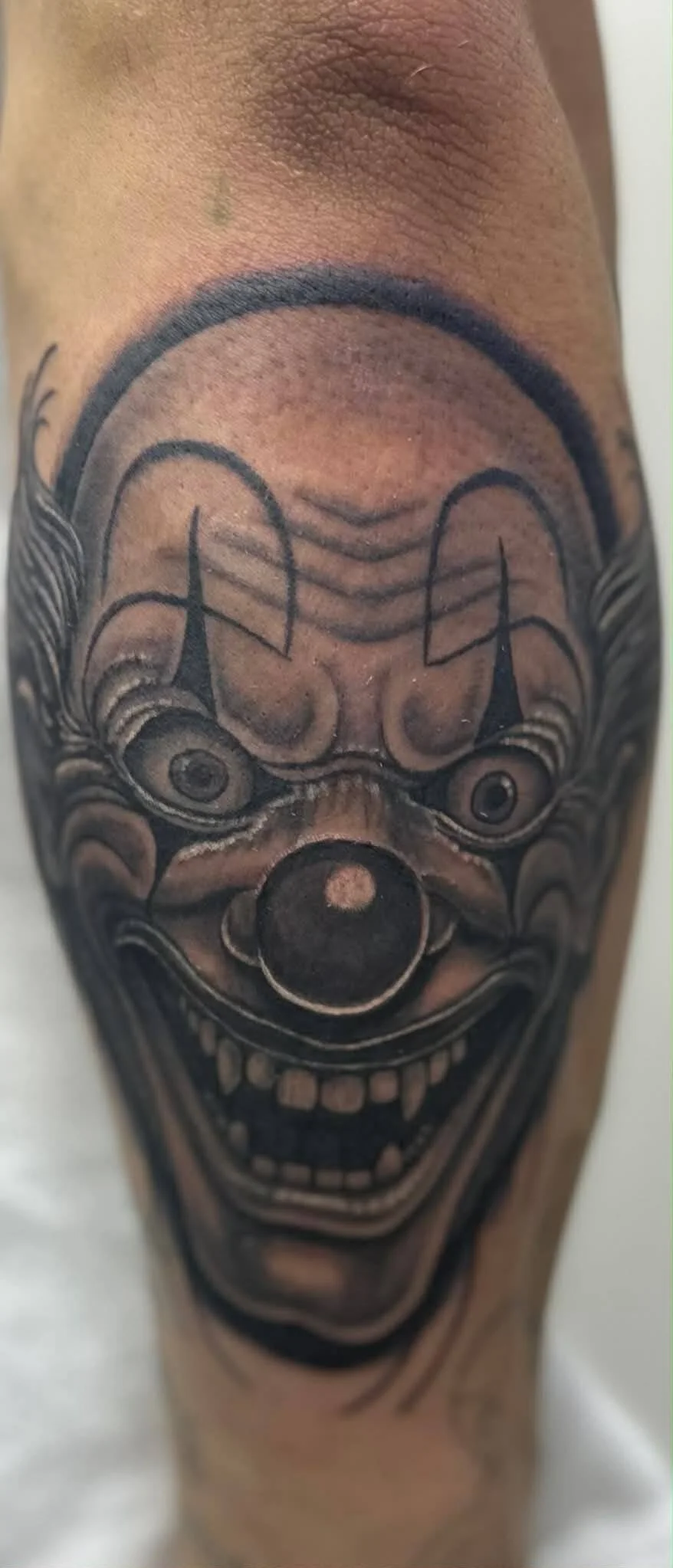 Tattoo of a clown face with a big smile, wide eyes, and a circular nose on a person's arm.