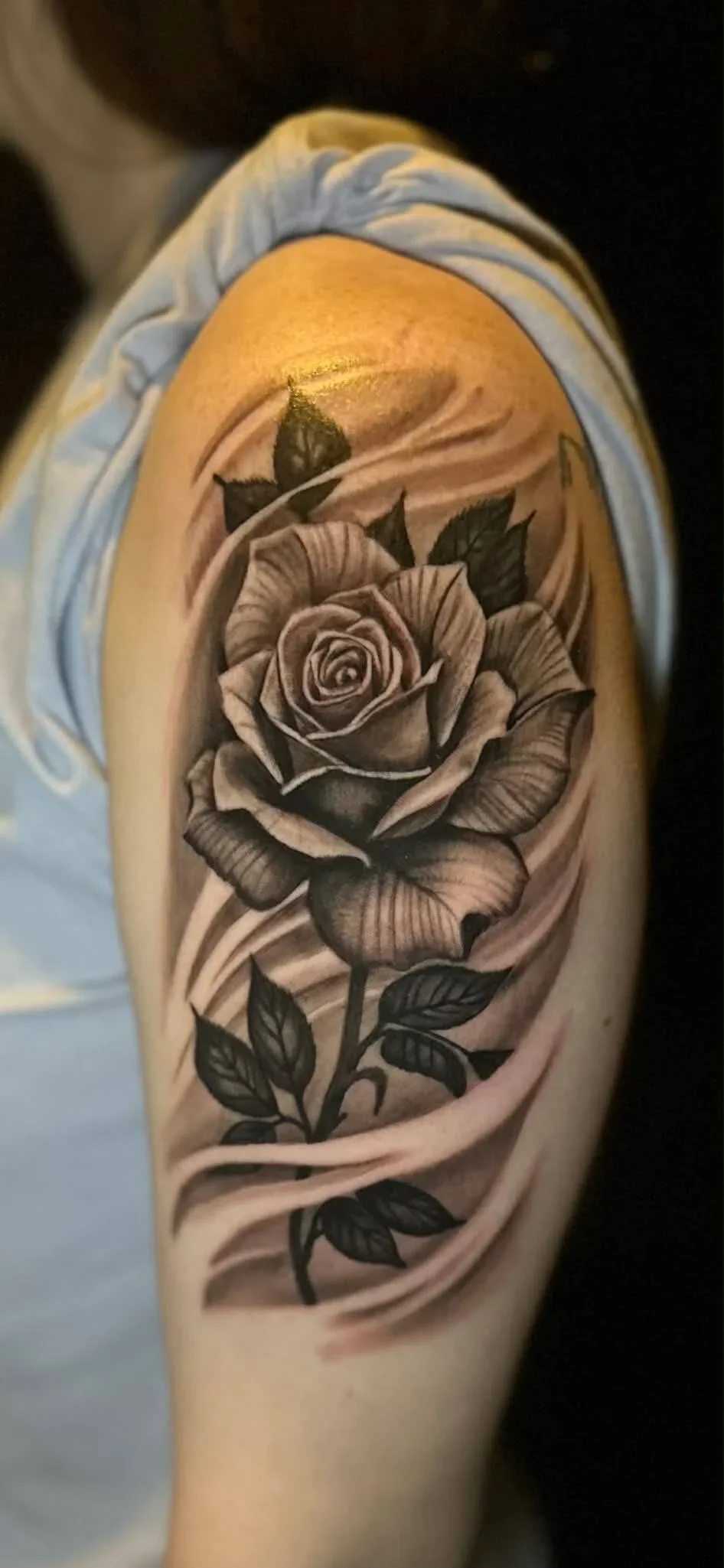 Black and gray tattoo of a realistic rose with leaves on a person's upper arm, with swirling shading in the background.