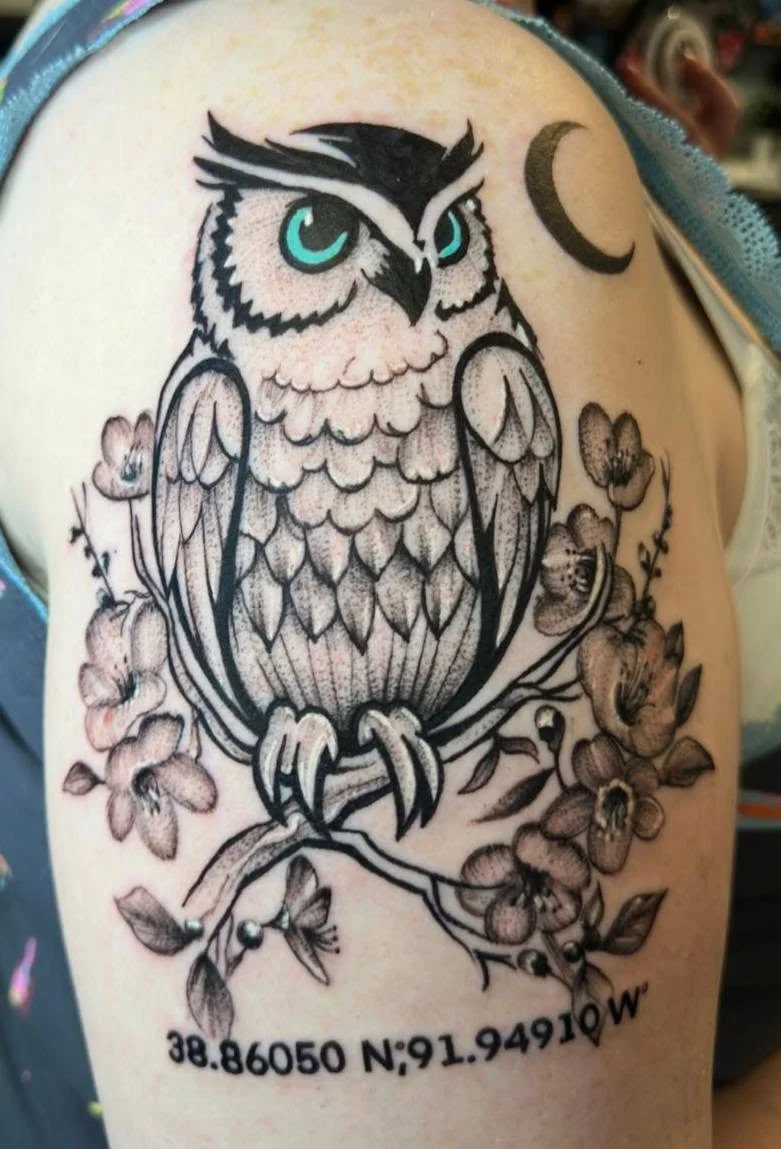 Tattoo of an owl with piercing teal eyes perched on a branch with flowers. The tattoo includes a crescent moon and geographic coordinates below.