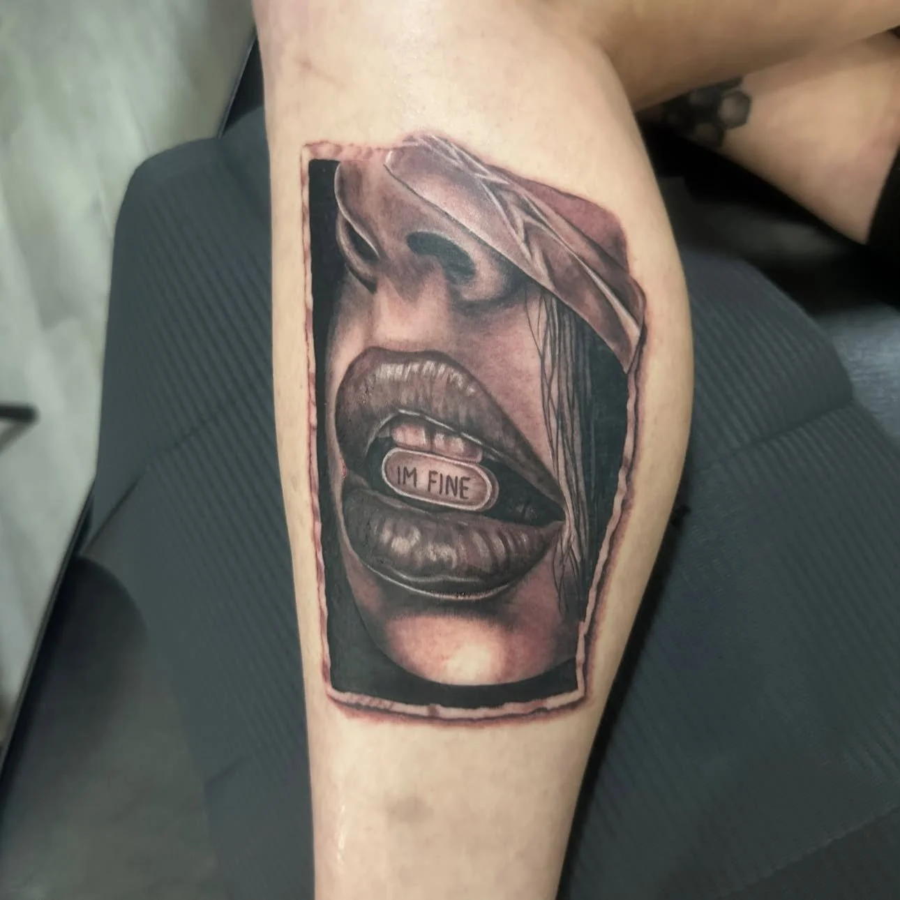 Realistic black and gray tattoo of a woman's face with an open mouth, showing a capsule labeled 'I'M FINE' inside her tongue.