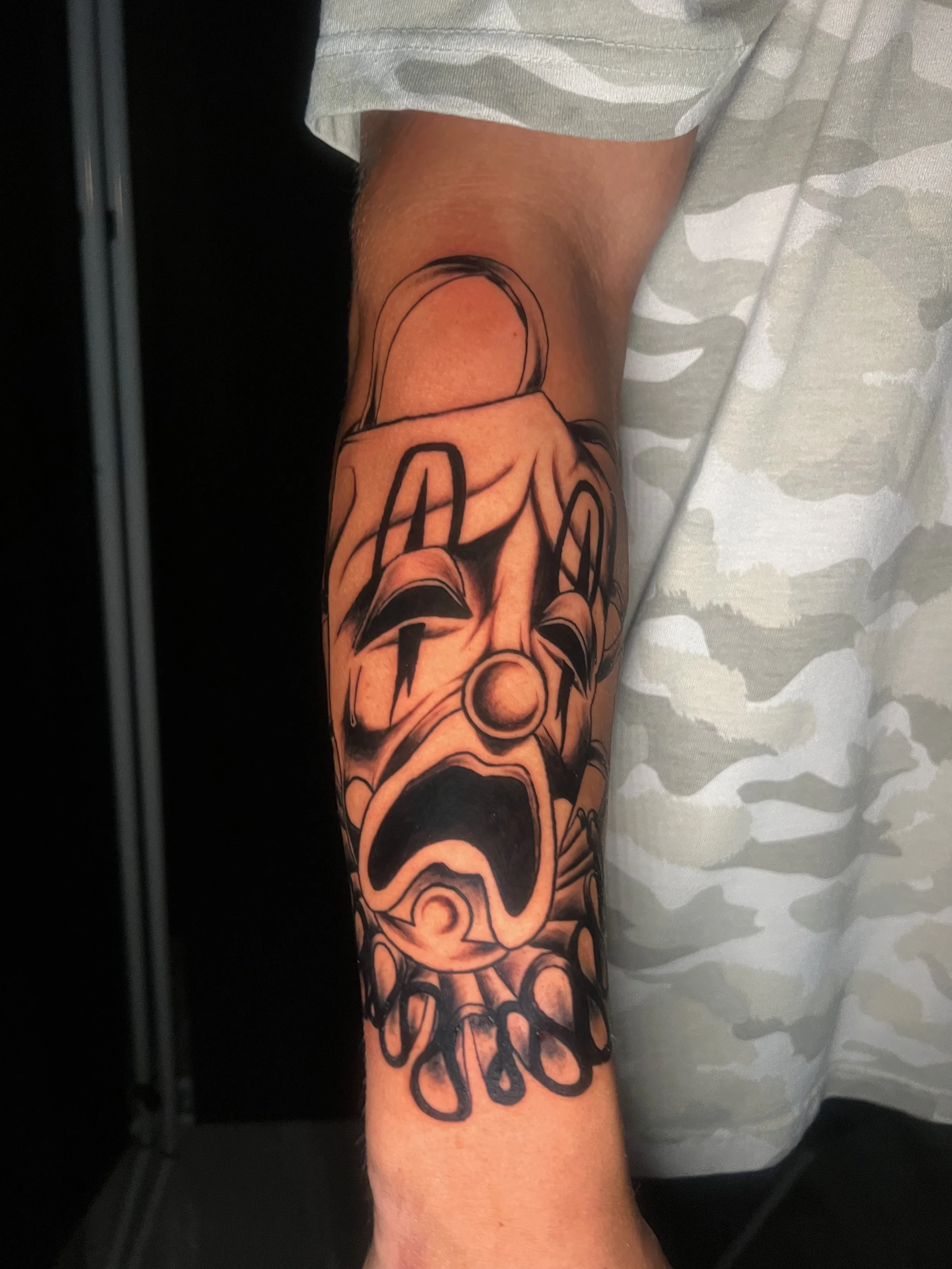 A tattoo of a skull on a person's forearm, with intricate black and gray shading, in front of a person wearing a light gray and white camo shirt.