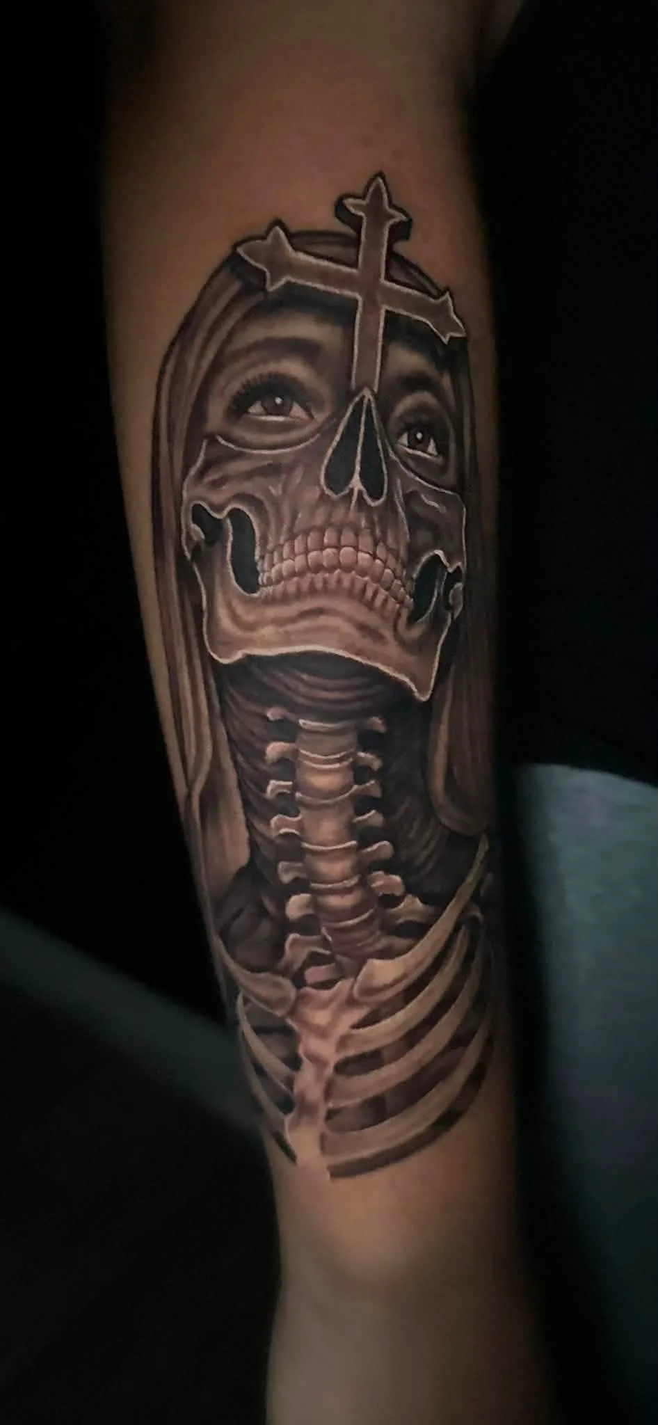 Tattoo of a skull with human eyes and a cross on top, combined with a skeleton's neck and ribcage, in black and gray tones.