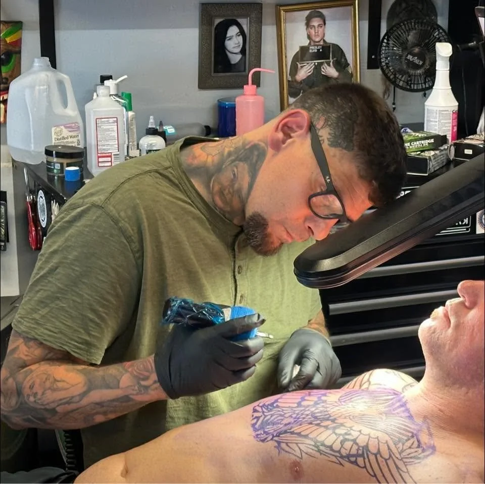 Tattoo artist in a green t-shirt and black gloves is tattooing a dragon design on a man's chest in a tattoo studio, with framed photographs on the wall behind him.