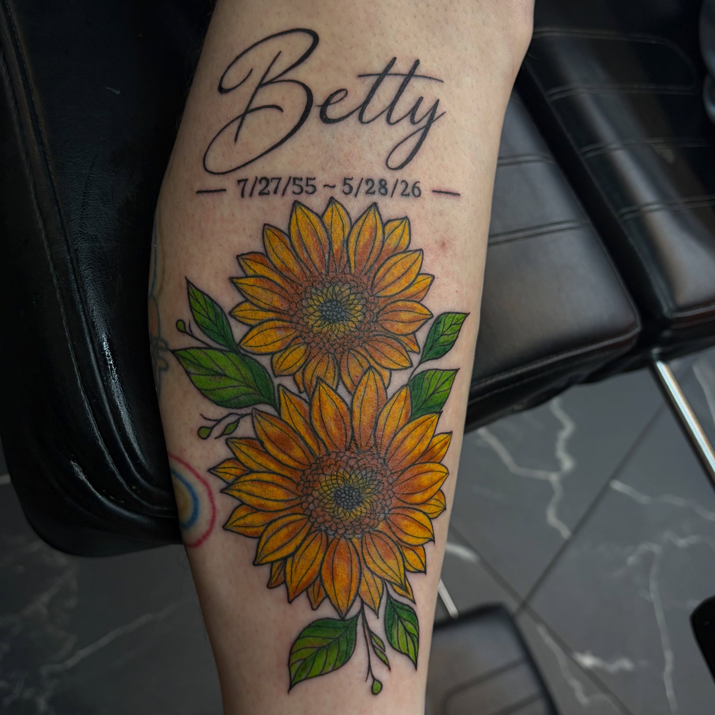 Tattoo on arm with the word 'Betty' in cursive and a sunflower with green leaves below it.