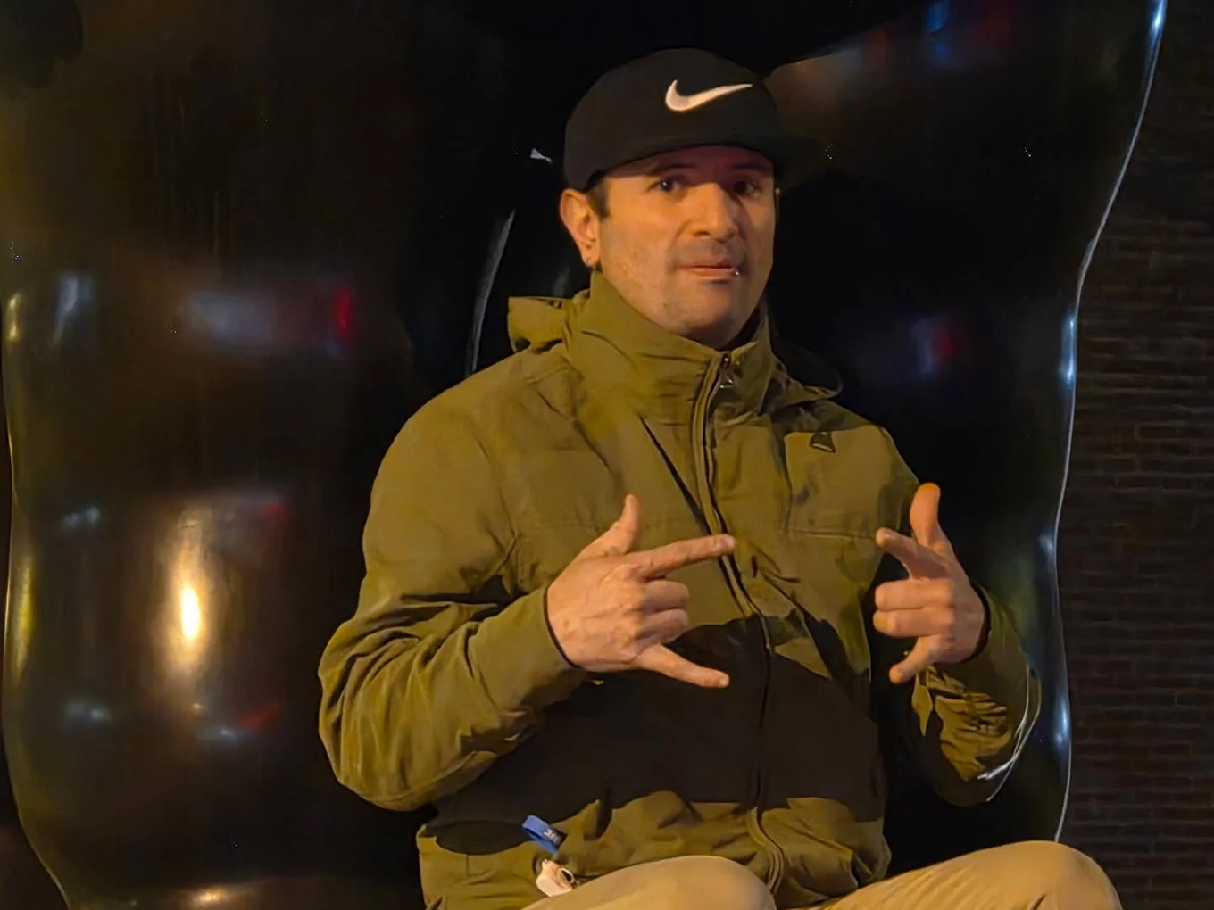 A man wearing a black Nike cap, brown jacket, and tan pants, making a rock-on gesture with both hands and sitting in front of a black reflective background.