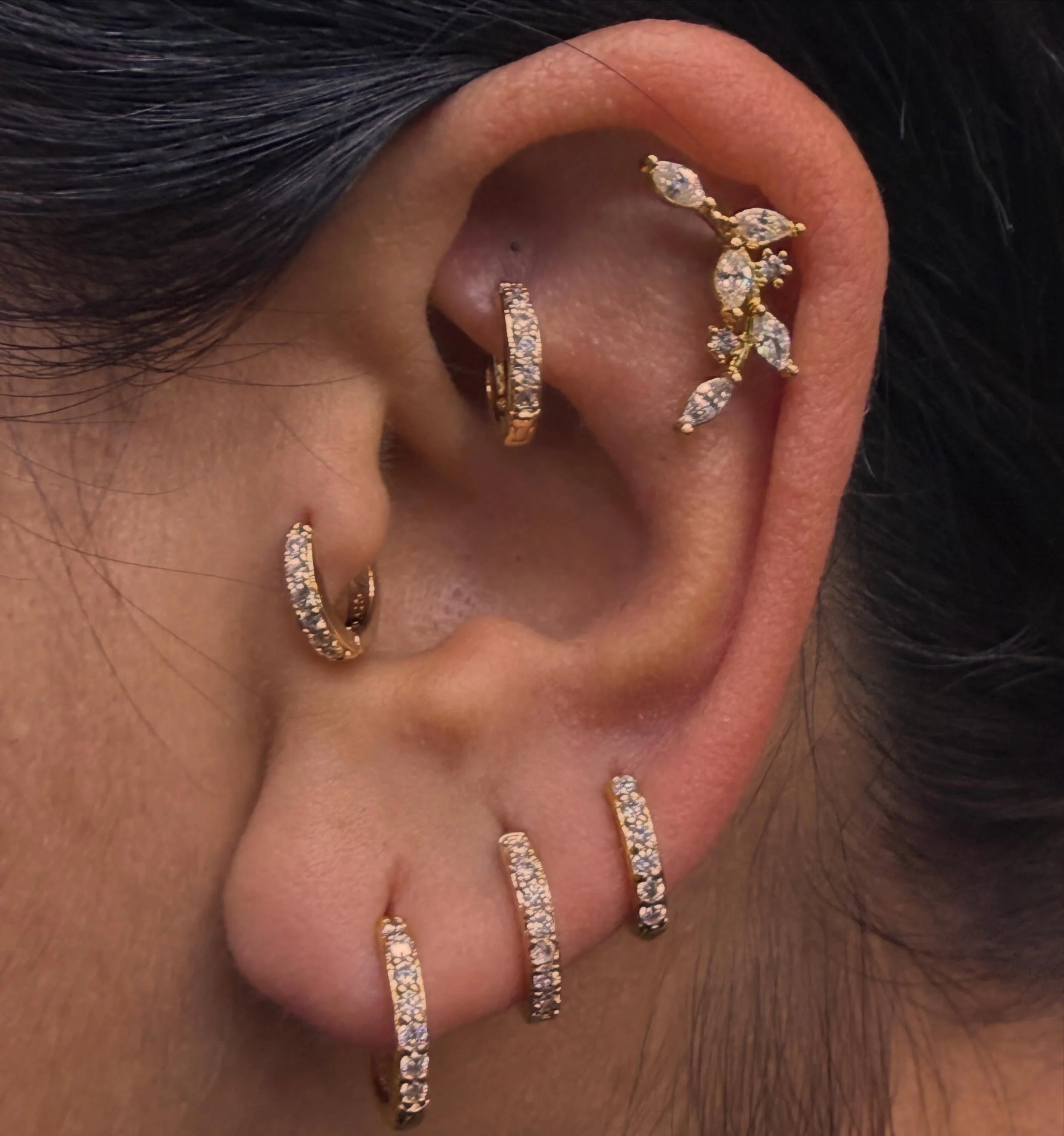 Close-up of a person's ear with multiple earrings, including a gold hoop in the earlobe, a gold cuff in the cartilage, a delicate chain with small decorative charms, and a bar-shaped earring with a patterned design, surrounded by dark hair.