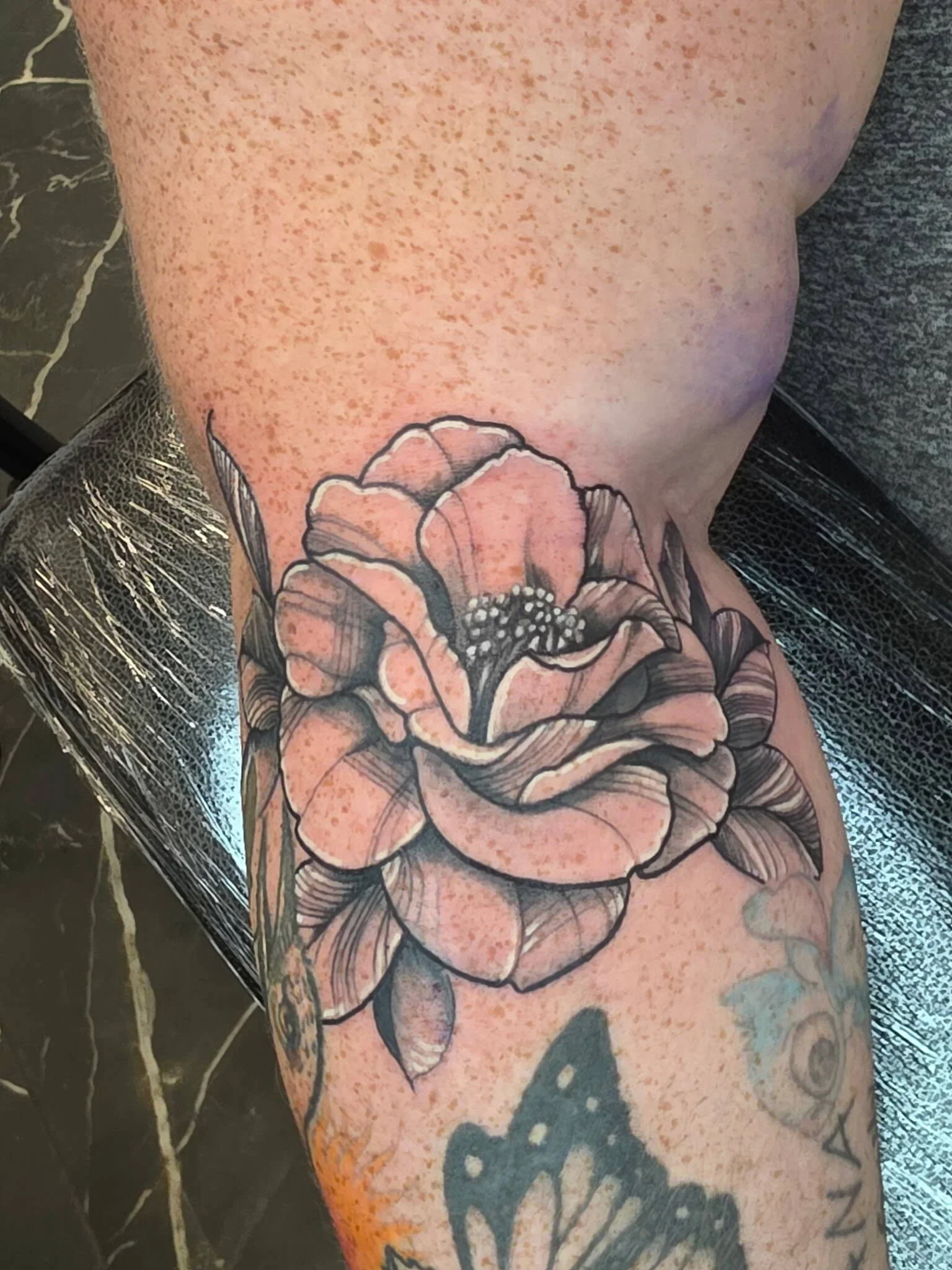 Close-up of a tattoo of a large flower with detailed petals on a person's arm, with a background of black and white marble flooring.