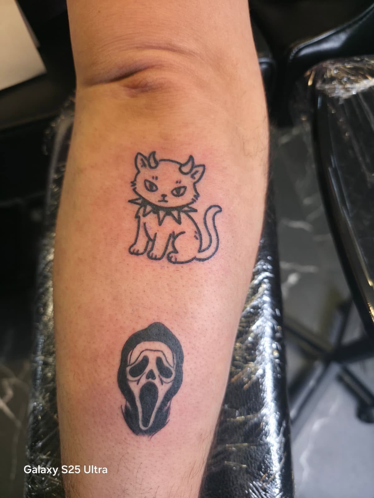Tattoo of a cute, cartoon-style, black outline cat with large eyes, small nose, pointy ears, and a curled tail, on a person's forearm.