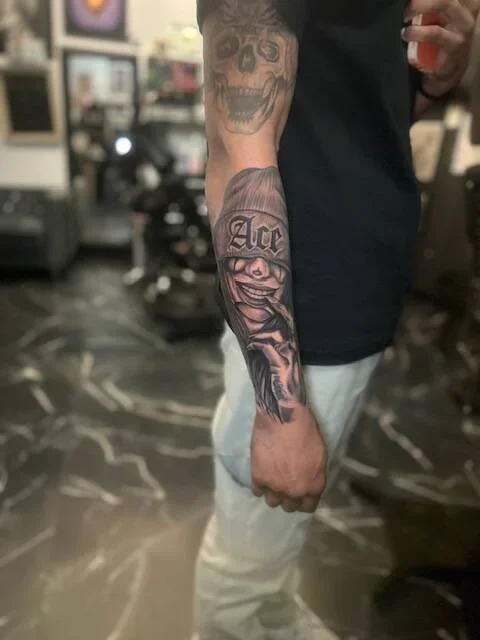 Tattooed arm with a skull on the upper arm and a clown face with the word 'Ace' on the lower arm.