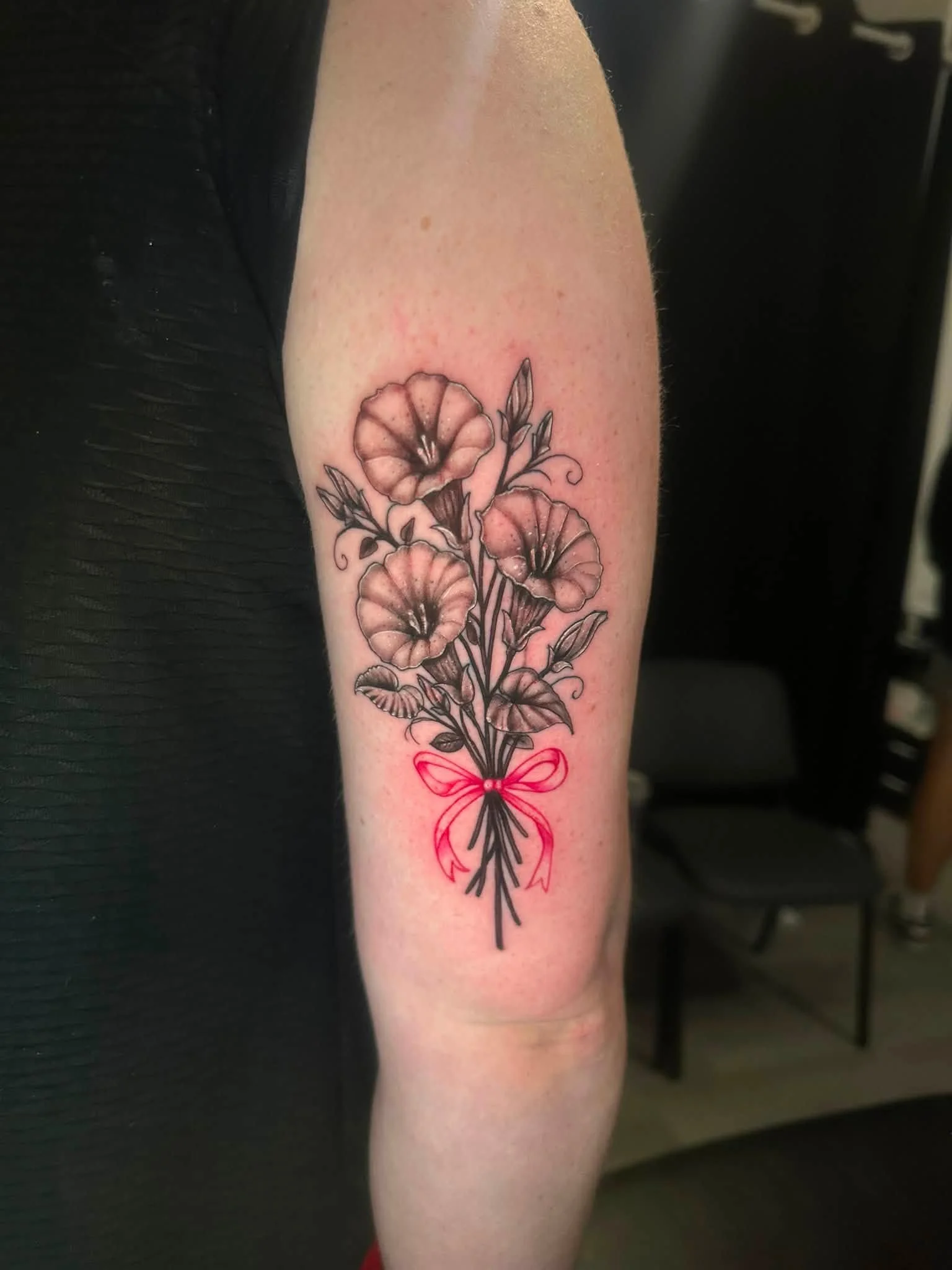 Black and gray floral tattoo on person's upper arm featuring three large flowers with detailed petals and leaves, decorated with a red bow at the bottom.