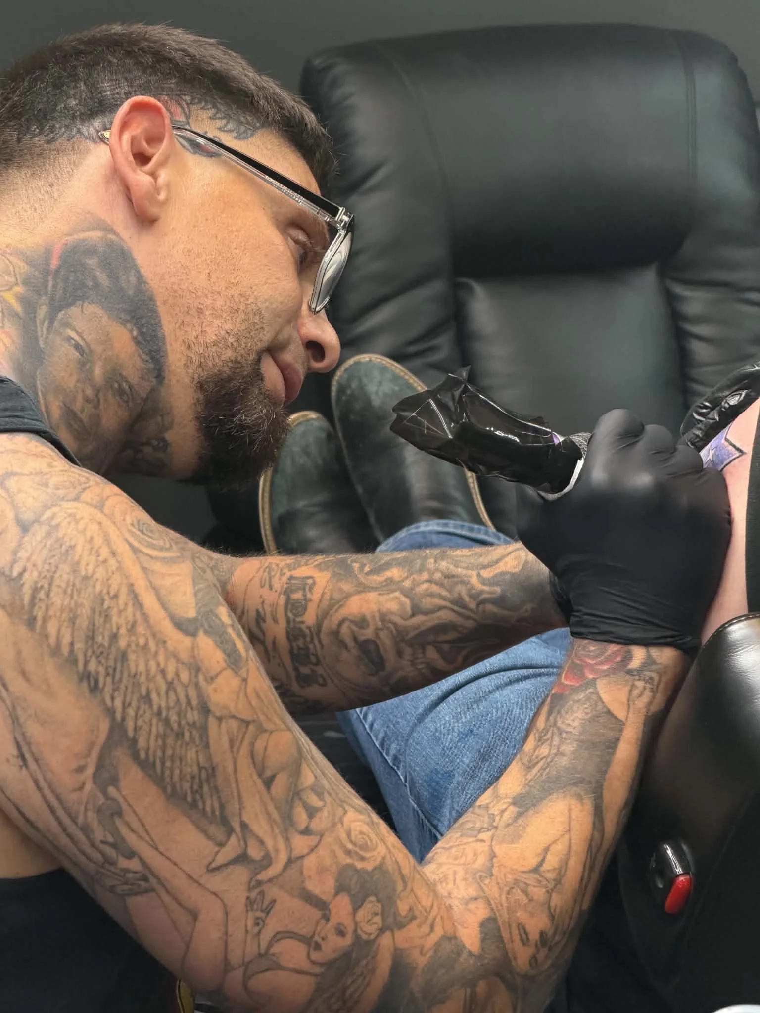A man with tattoos on his arms and face, wearing glasses, is sitting in a black leather chair while getting a tattoo on his arm.