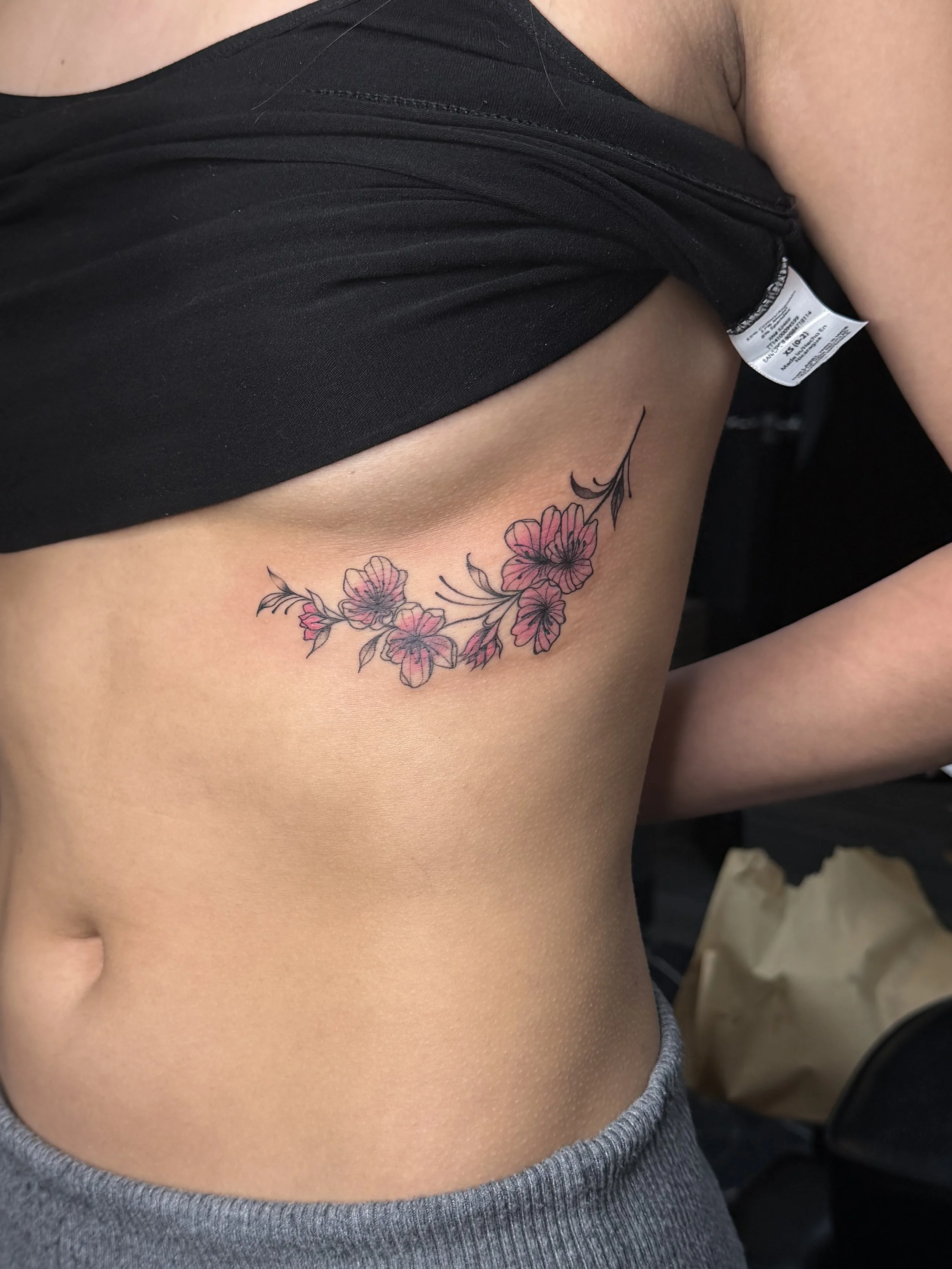Close-up of a person's side showing a floral tattoo on their ribs, partially covered by a black shirt and grey pants.
