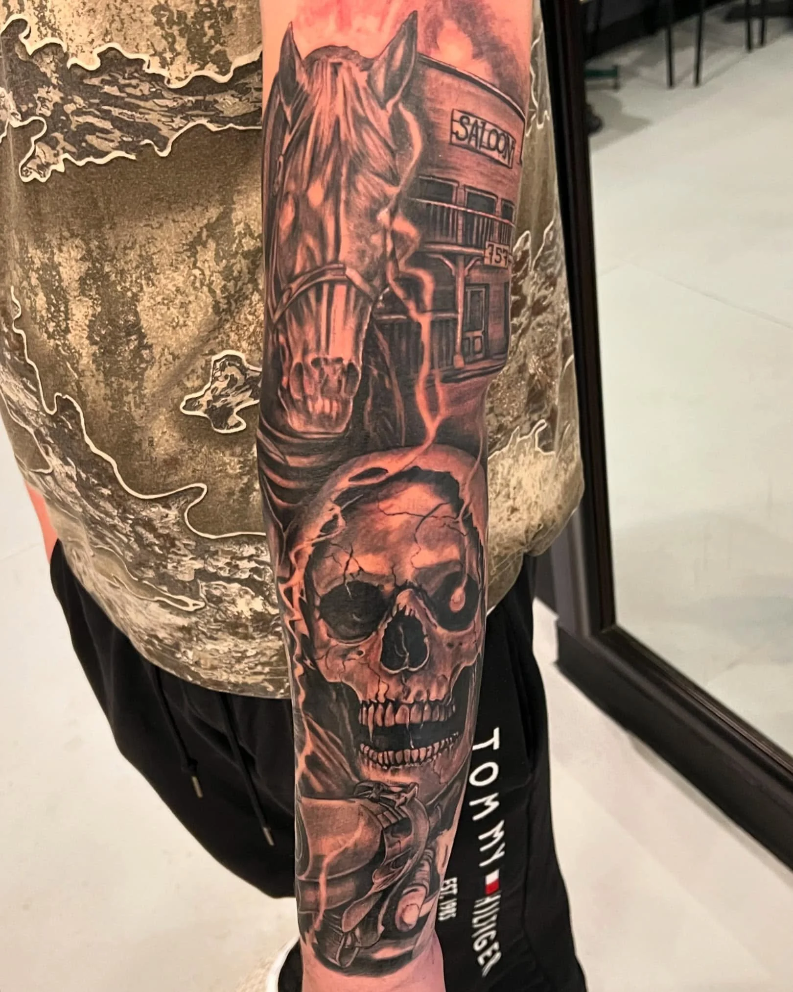 A person's arm with a tattoo of a skull and a building labeled 'Saloon' with a bus in front of it.