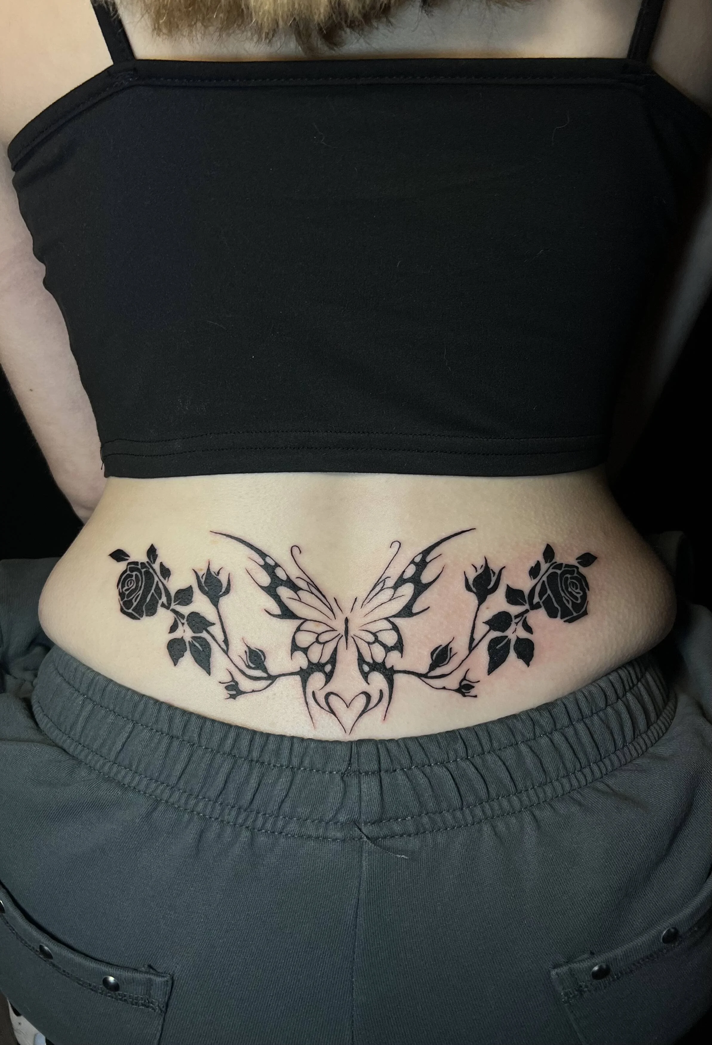A tattoo on a person's lower abdomen depicting a symmetrical design of a butterfly in the center, with roses and leaves on either side, and a heart shape at the bottom.