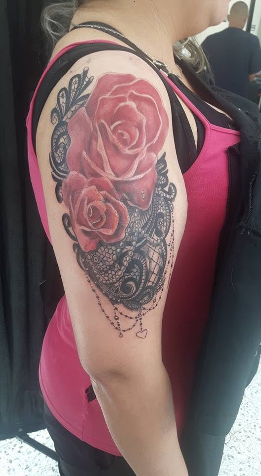 Tattoo of two large roses with dark ornamental lace and beaded jewelry details on the upper arm of a woman wearing a pink tank top and black outerwear.