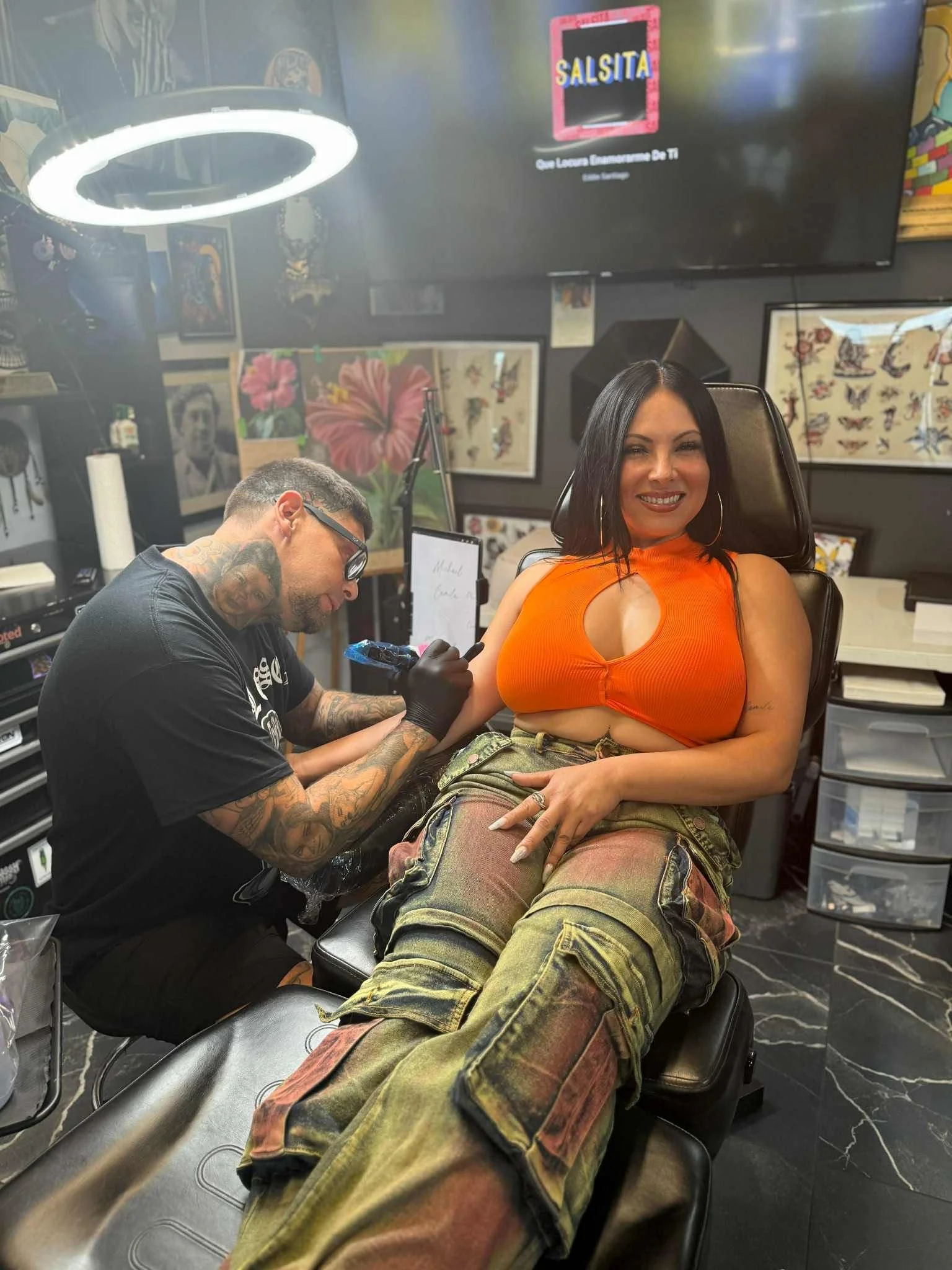 A woman smiles while getting a tattoo on her upper arm by a tattoo artist in a tattoo studio. The woman is wearing an orange top and camouflage pants. The studio has various artwork on the walls and studio equipment around.
