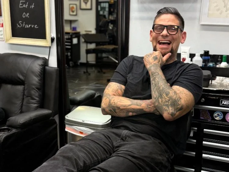Man with tattoos and sunglasses standing outside tattoo shop, wearing a black tank top and holding sunglasses in his right hand.