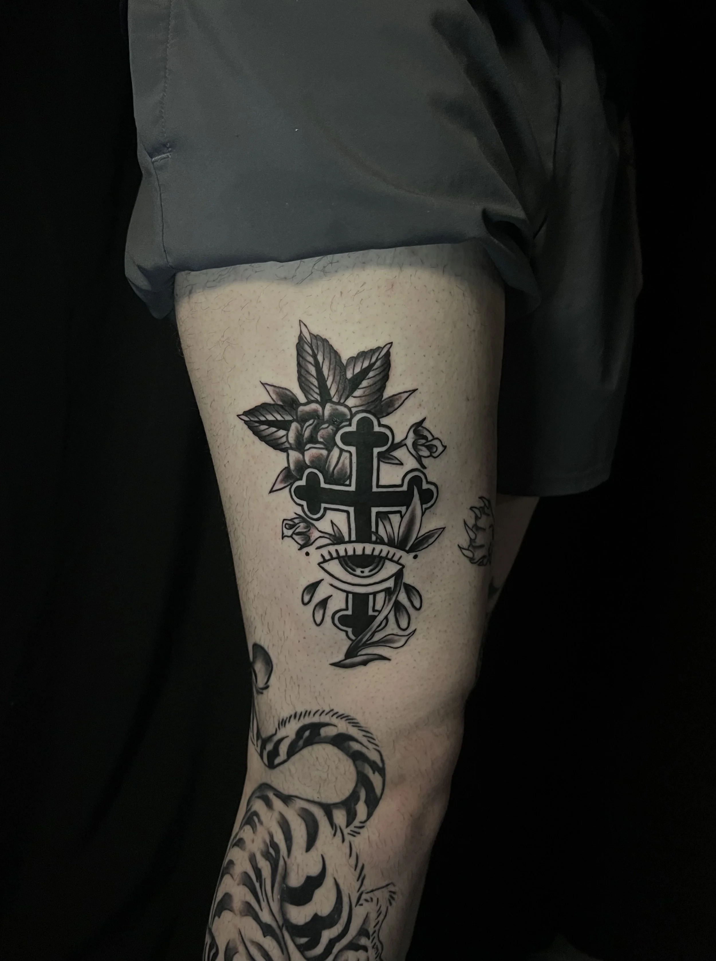 Close-up of a person's thigh with a black tattoo of a tiger and a medical cross with an eye and leaves. The person is wearing dark shorts.