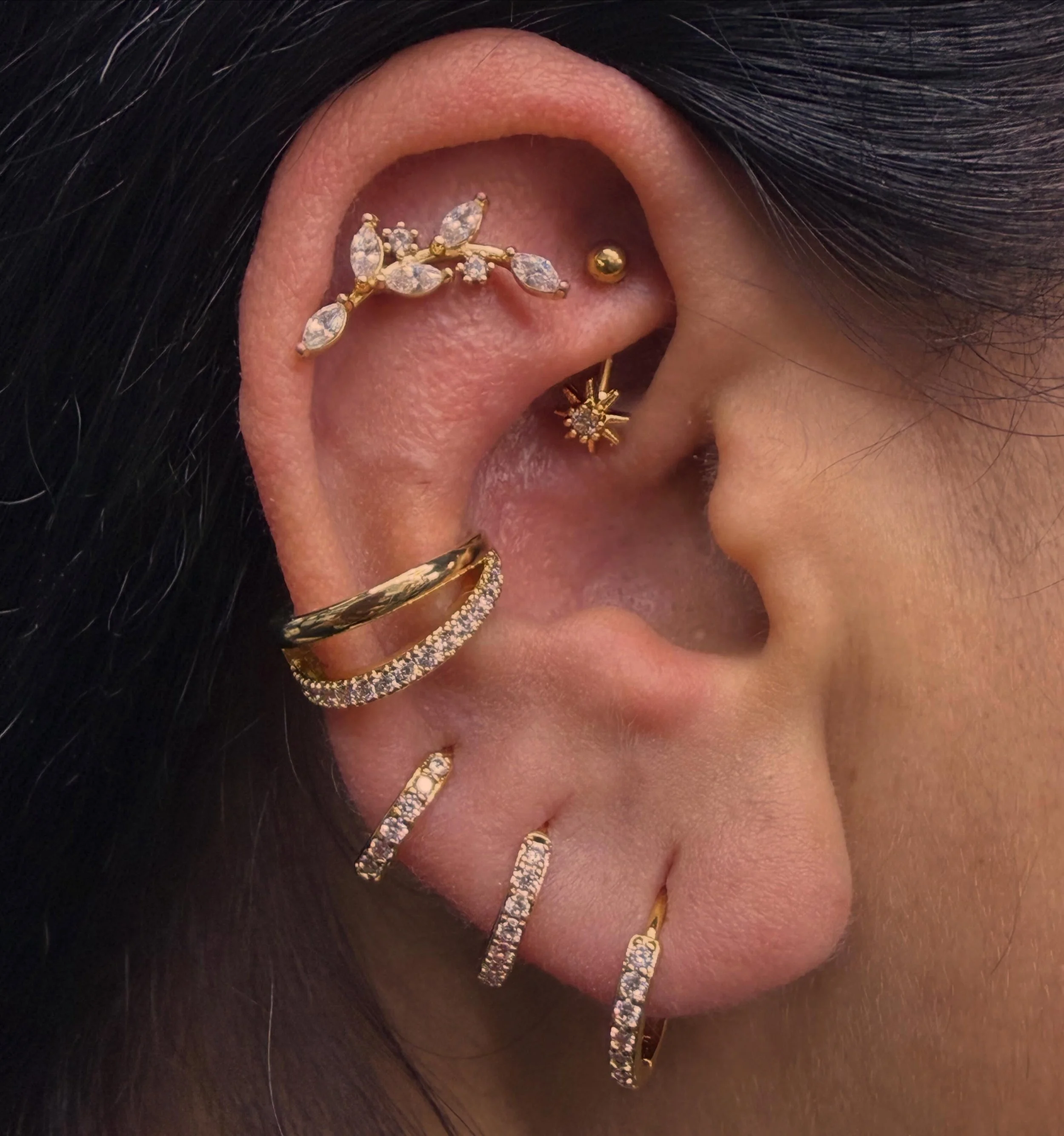 Close-up of a person's ear with multiple gold and diamond earrings, including a hoop earring with diamonds, a star-shaped earring, and several small studs, set against dark hair and skin.