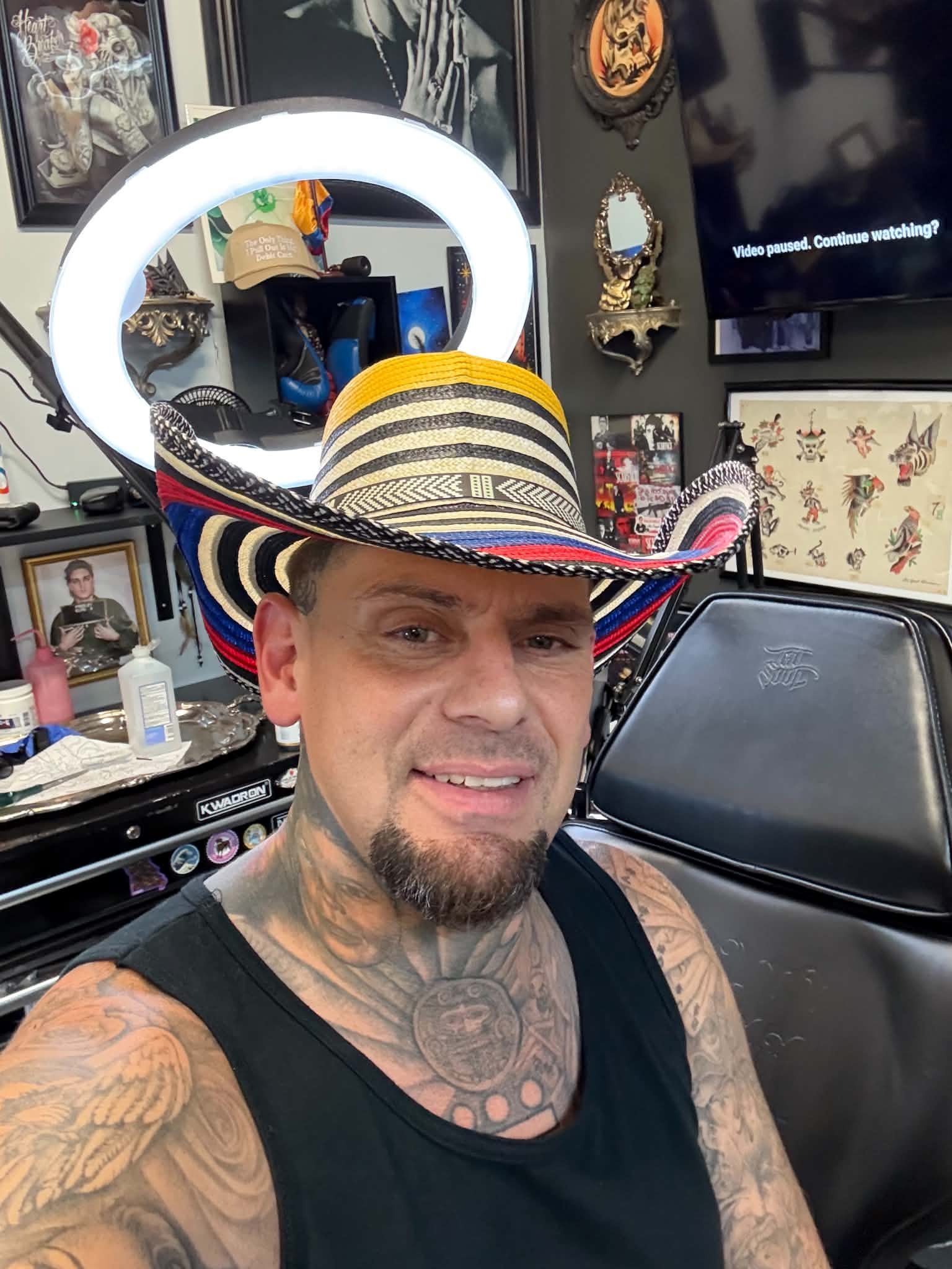 A man with tattoos on his arms and chest wearing a black tank top and a large multicolored sombrero, taking a selfie in a room with tattoos and artwork on the walls, a television paused, and various items on a nearby table.