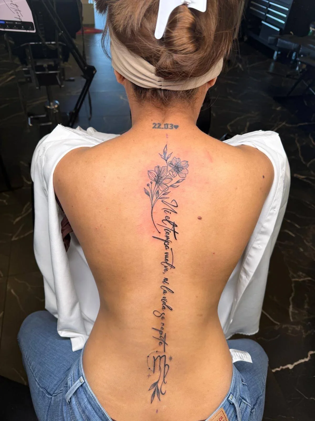 A person with a floral tattoo on their back that includes flowers and cursive script. The person is sitting, with their back facing the camera, wearing a white shirt and a beige headband, with a tattoo machine nearby.