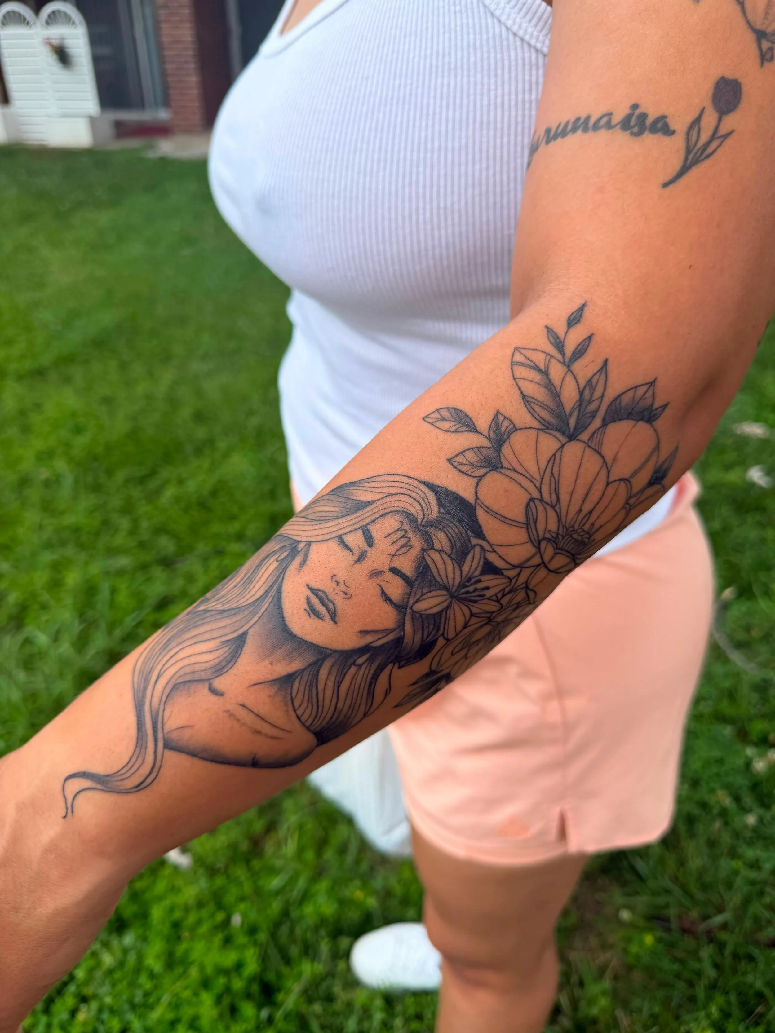 Close-up of a person's arm with tattoos, including a woman's face, flowers, and a quote in cursive writing. The person is wearing a white sleeveless top and pink shorts, standing outdoors on green grass.