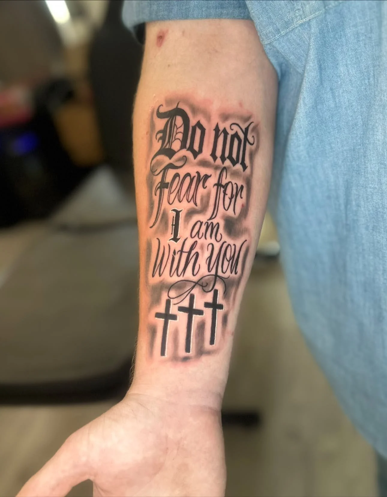 Tattoo on person's forearm with the quote 'Do not fear for I am with you' in gothic and cursive fonts, along with three crosses below.