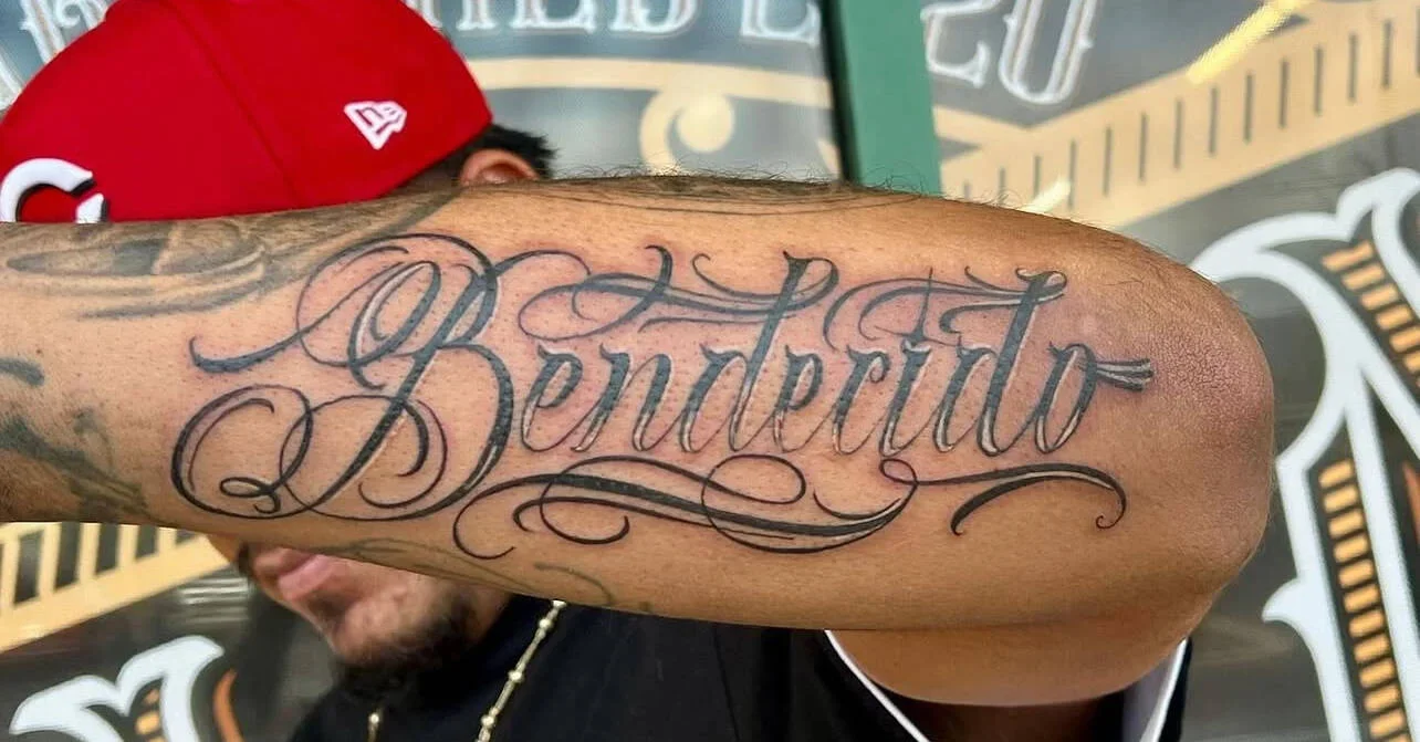 Tattoo of the word "Bentevette" in elaborate black script on a man's upper arm, with a man wearing a red cap in the background.