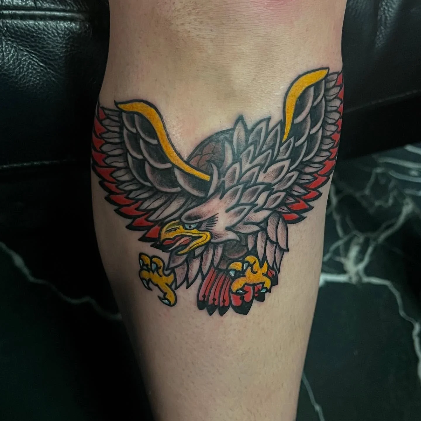 Tattoo of a bold eagle with outstretched wings in black, gray, red, yellow, and orange colors on a person's upper arm.