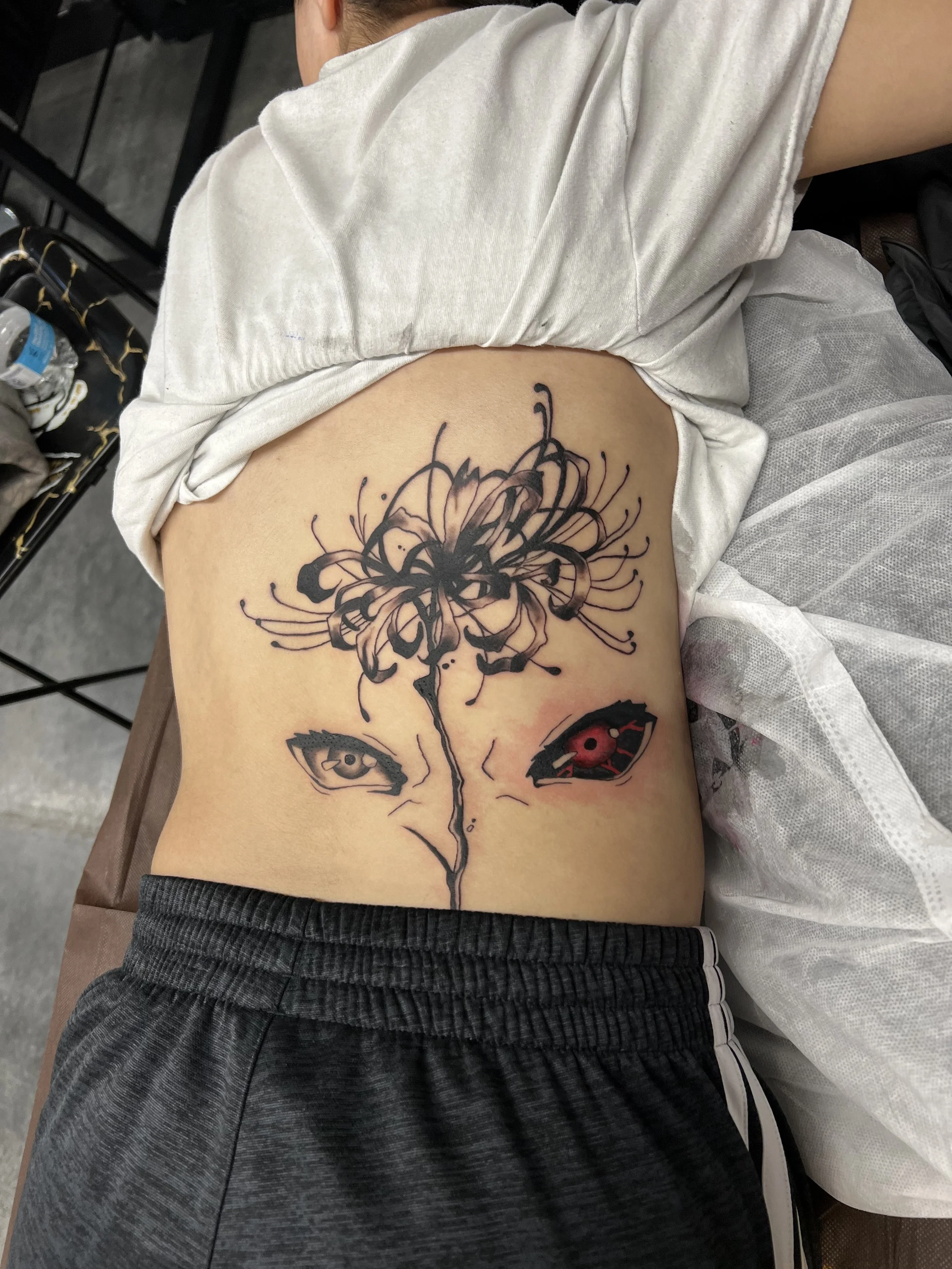 A tattoo of a flower with intricate black ink design on a person's side ribcage, featuring a butterfly-like face with red eyes.