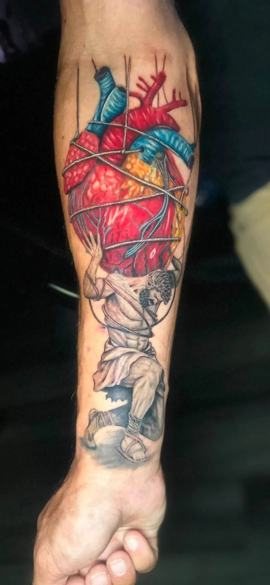 Tattoo of a person carrying a large, detailed anatomical heart with blue and red sections, wrapped with wire, and a man with a lion's head holding a spear at the bottom.